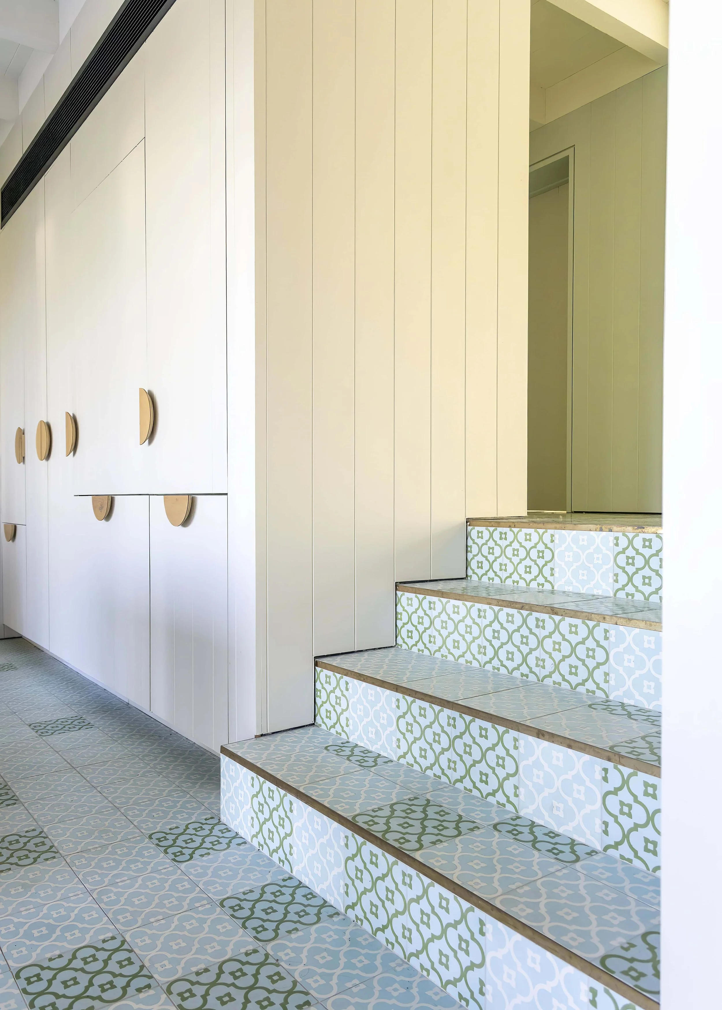Interior space with a pathway, a wall of white cabinets with wooden handles, and stairs tiled with patterned tiles in white and green. The steps have a modern design with a slight overhang.