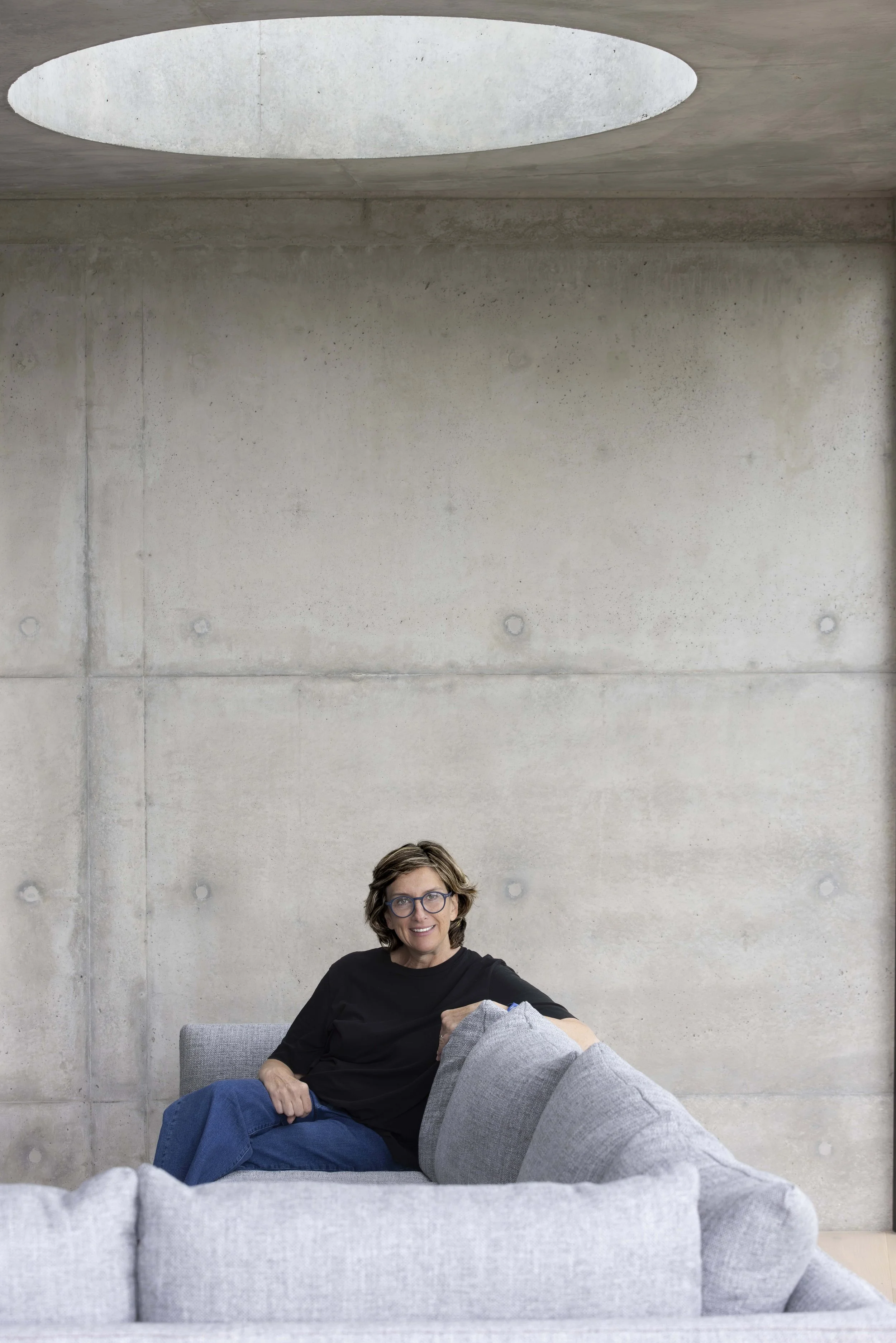 Woman with glasses sitting on gray couch in front of a concrete wall with a circular skylight above.