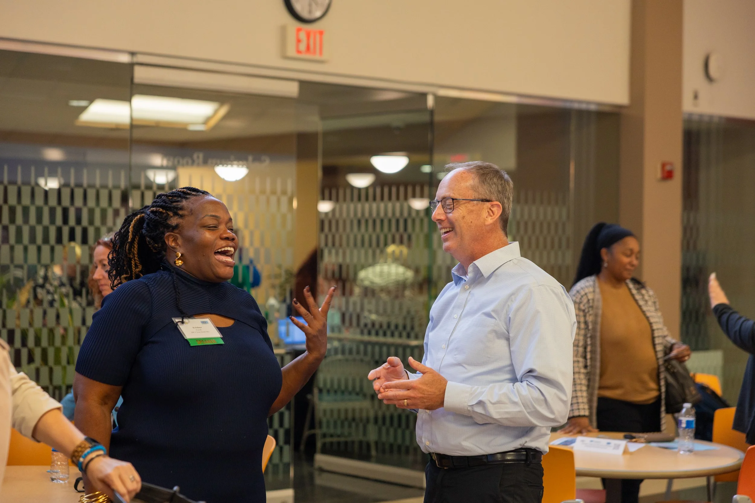 Two people, a woman and a man, are smiling and talking to each other in a lively conversation at an indoor event. Other people are visible in the background.