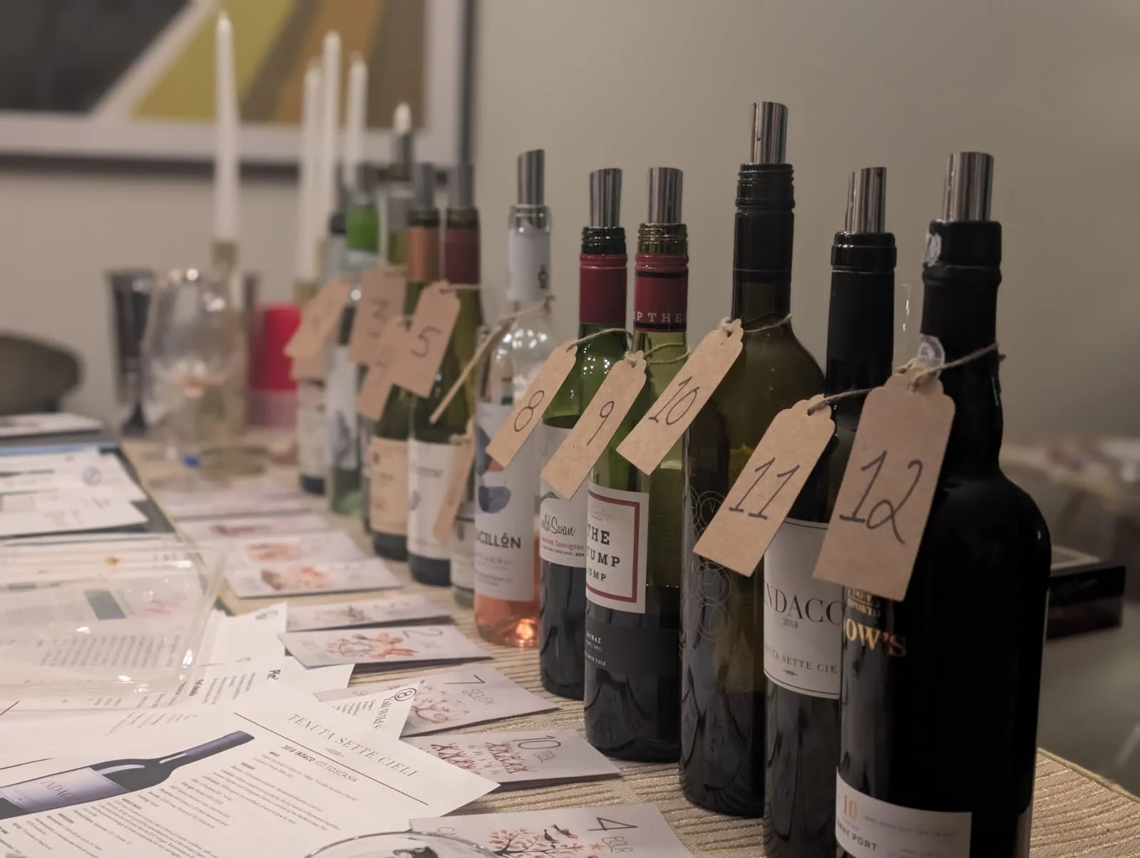 A row of wine bottles with numbered tags hanging from each, displayed on a table with scattered papers and informational flyers.