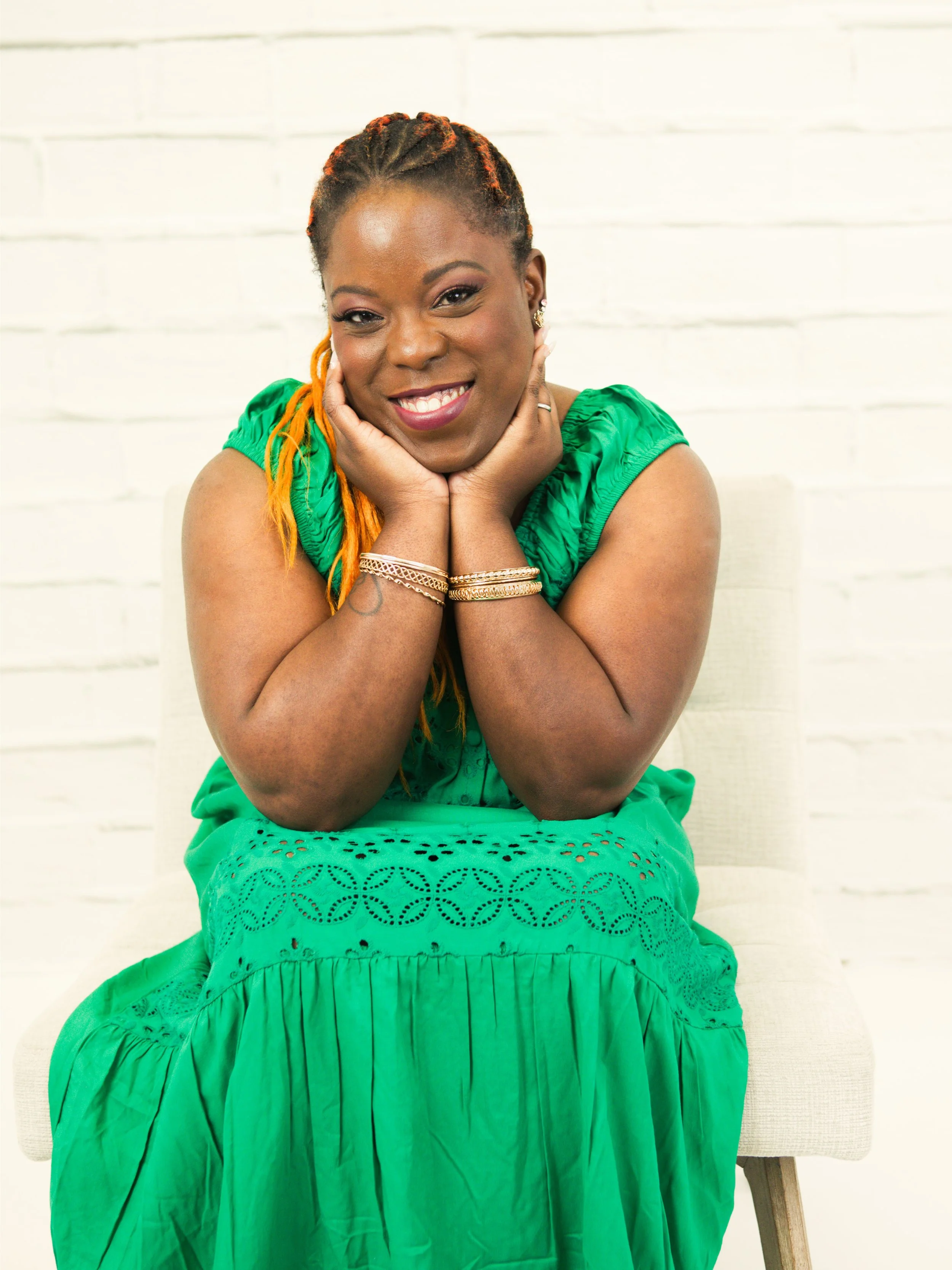 A woman in a green dress sitting on a cream-colored chair with a white brick wall background. She has braided hair with orange highlights, is smiling, and resting her chin on her hands.