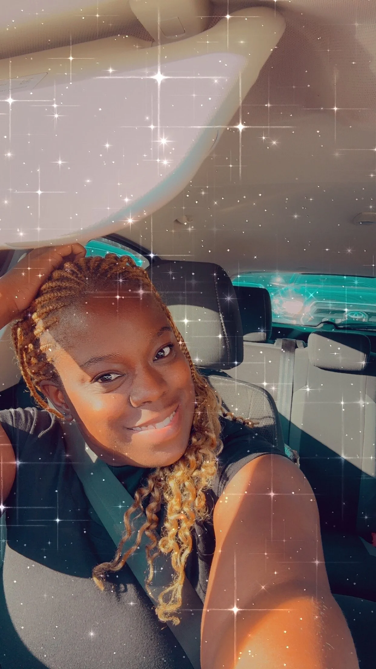 A young woman with braided blonde hair, smiling and sitting inside a car, taking a selfie with a filter that adds sparkling star effects.