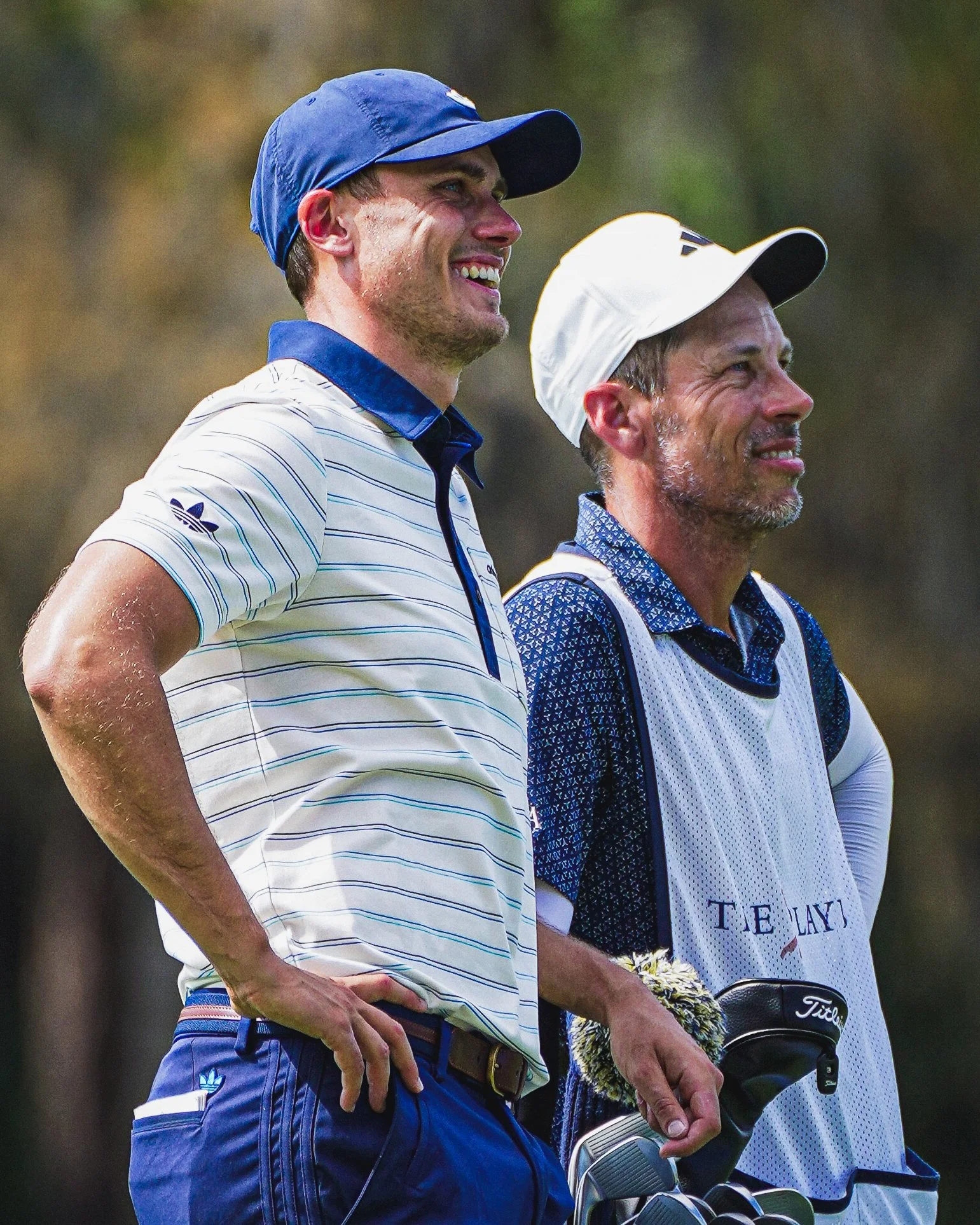 A woman shooting baskets in a basketball arena, dressed in a dark sports jacket and navy pants with an orange shirt underneath, surrounded by other players and spectators.