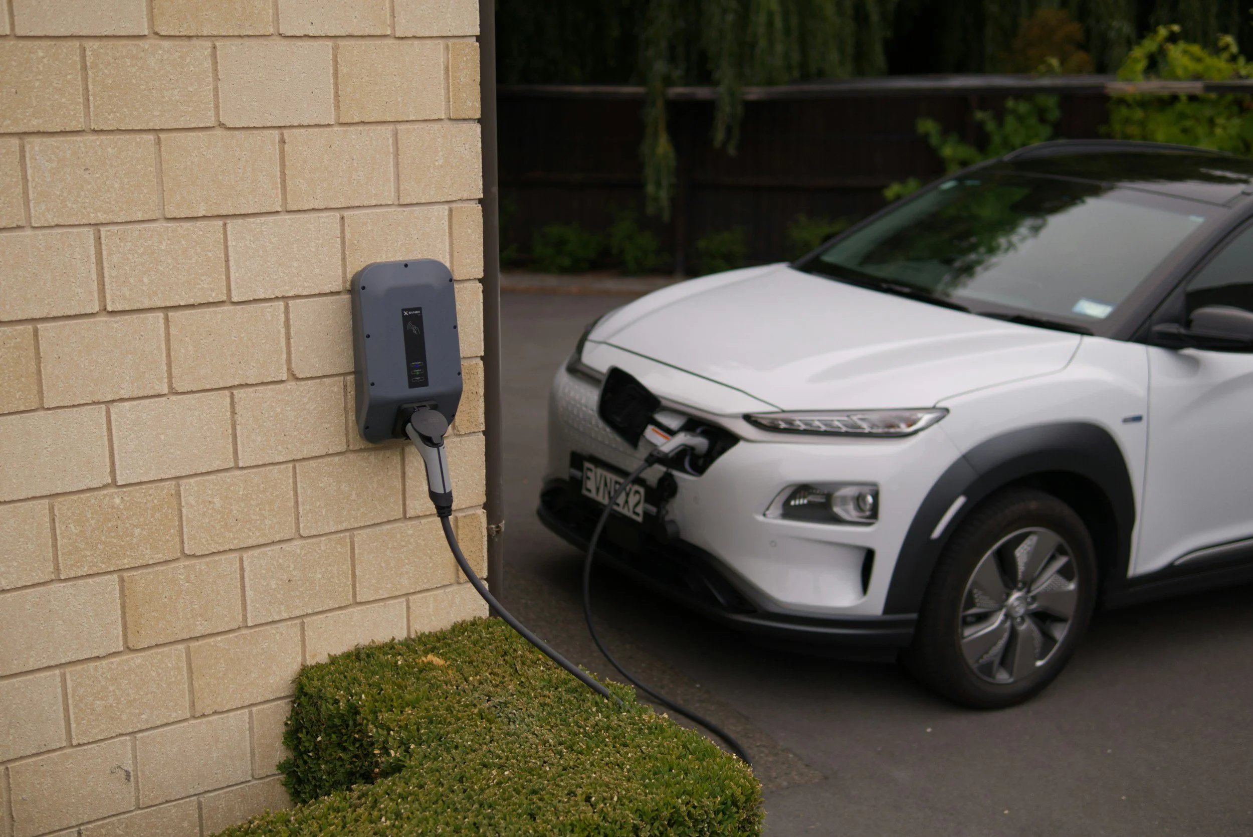 Electric vehicle charging station mounted on a beige brick wall with a white electric car plugged in for charging in a parking lot, with trees and a dark wooden fence in the background.