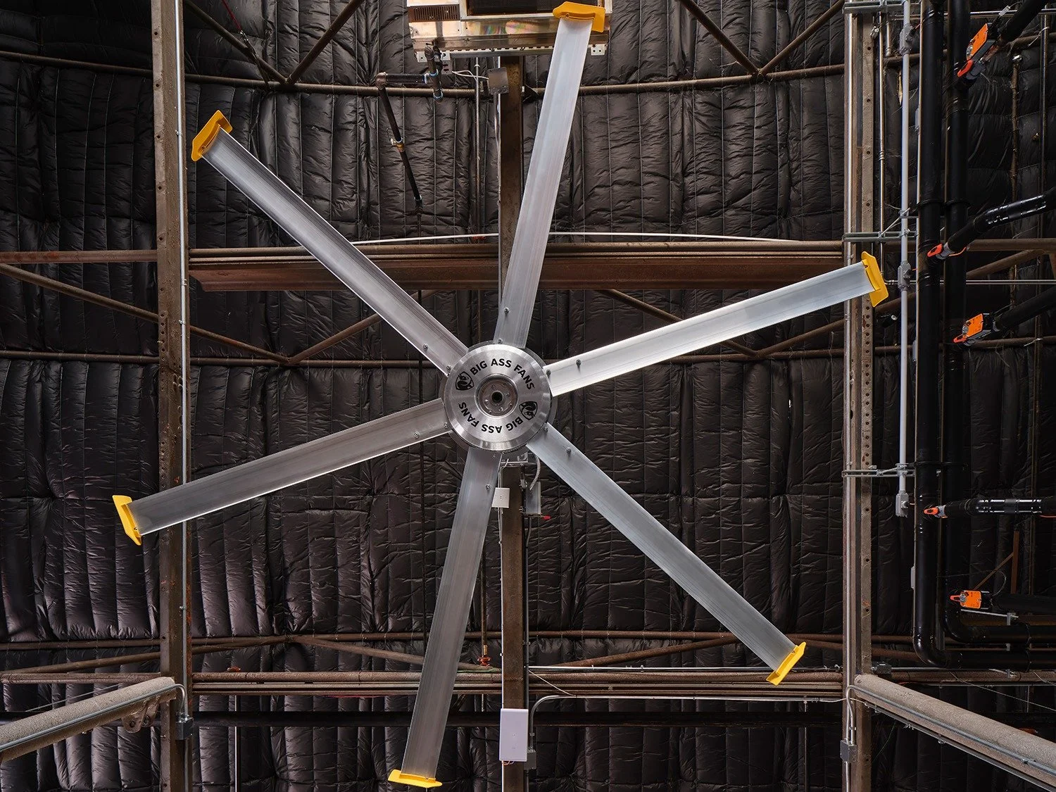 An industrial ceiling fan with six metallic blades and yellow tips, installed on a ceiling with black insulation and metal framing.