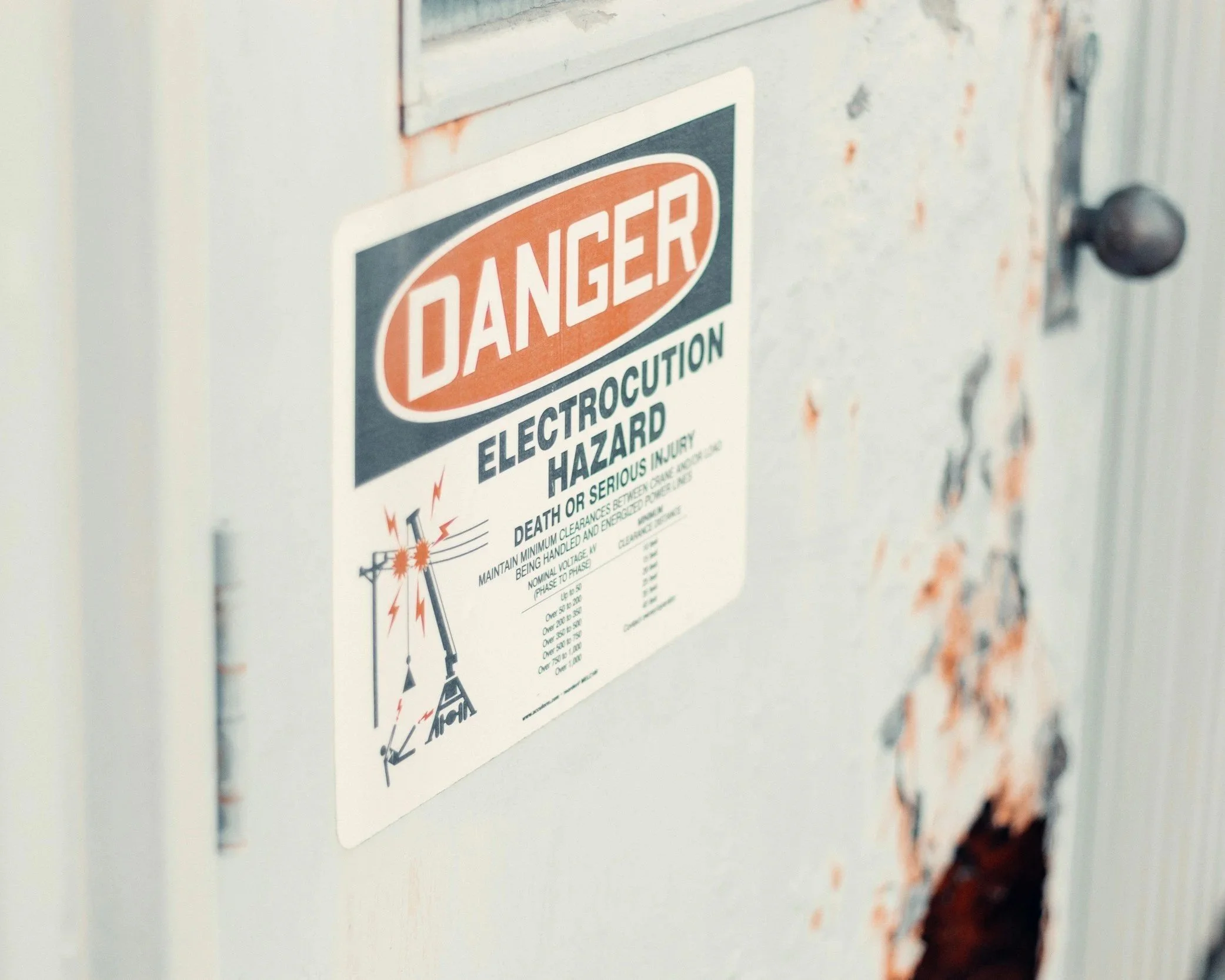 A warning label on a metal electrical box indicating danger of electrocution, with text warning of serious injury or death and a diagram of a power pole and electrical wiring.
