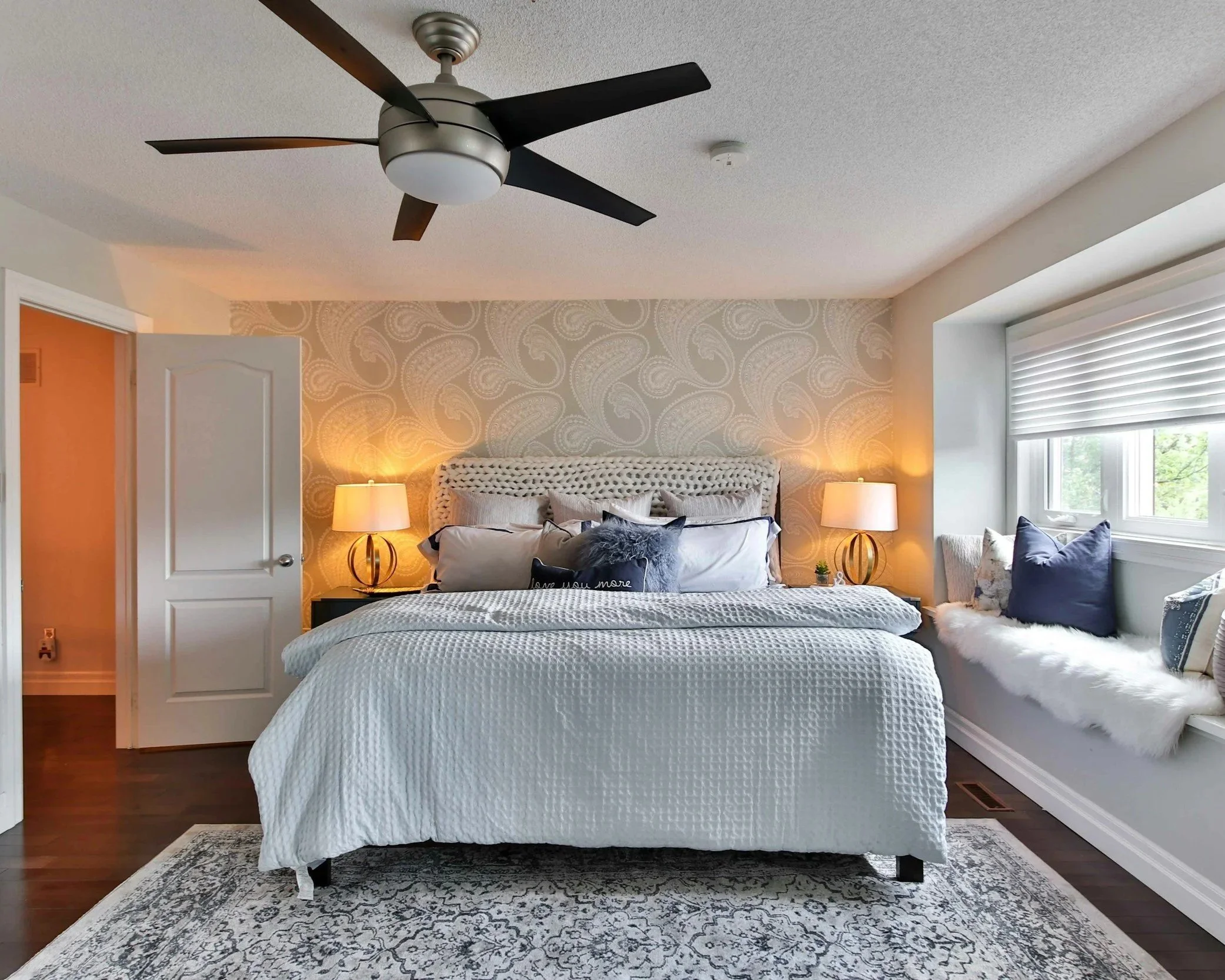A bedroom with a white bed, two bedside lamps, a window with blinds, a ceiling fan, and decorative pillows