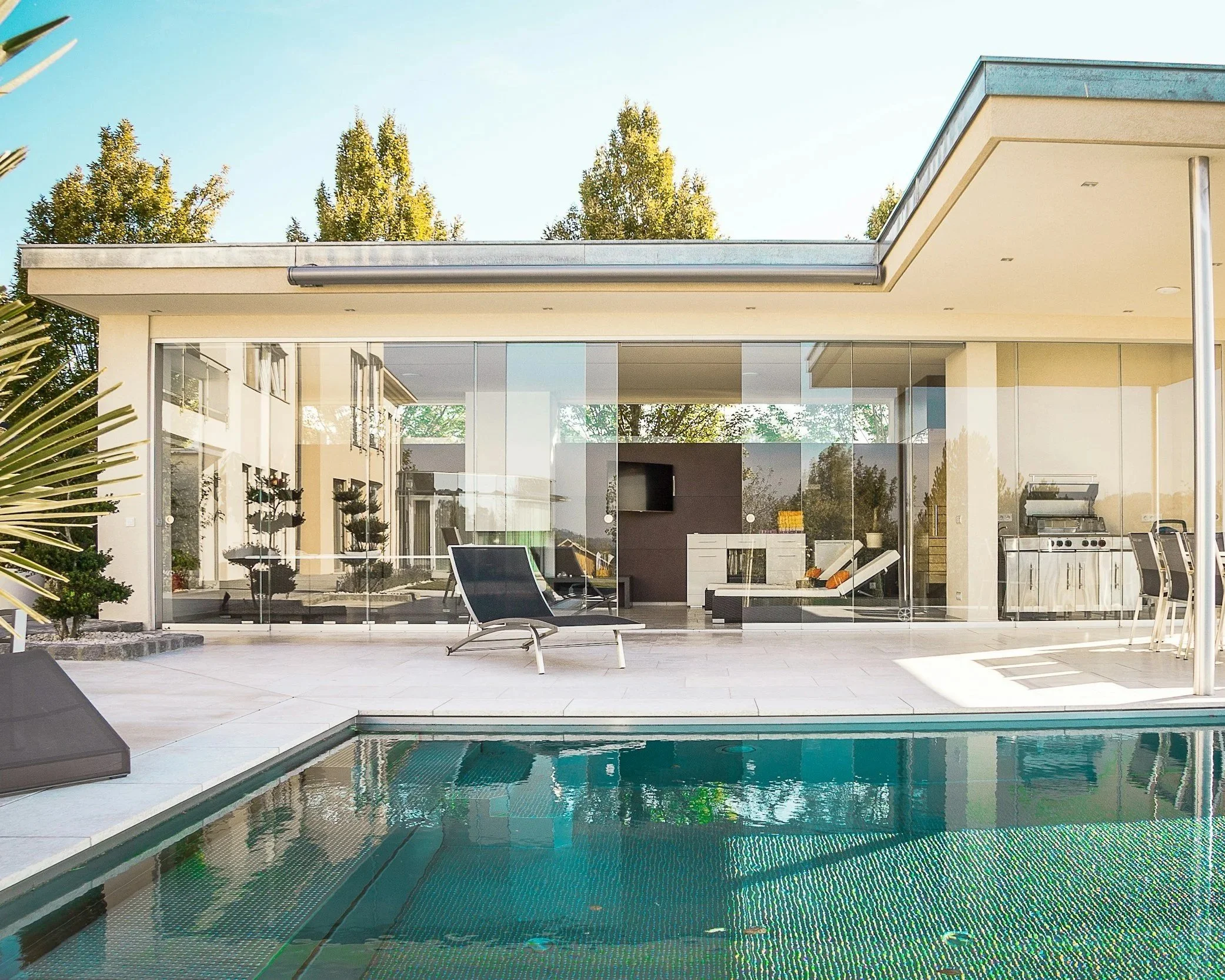 Modern house with glass walls, outdoor pool, and patio furniture, surrounded by trees.