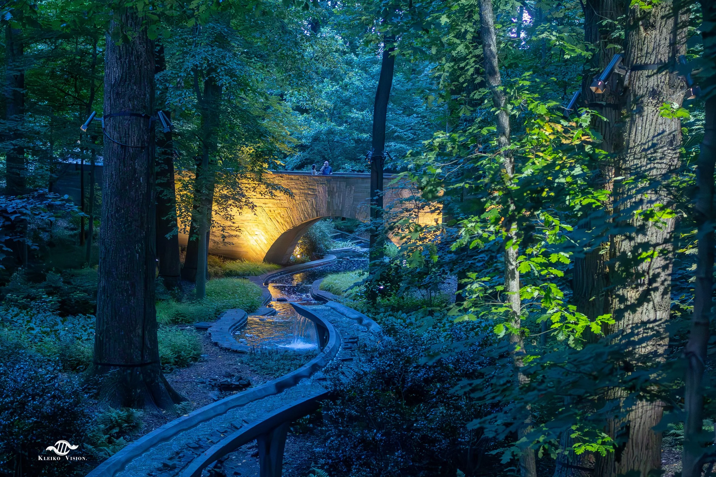 Illuminated stone bridge over a garden stream at dusk with two visitors crossing