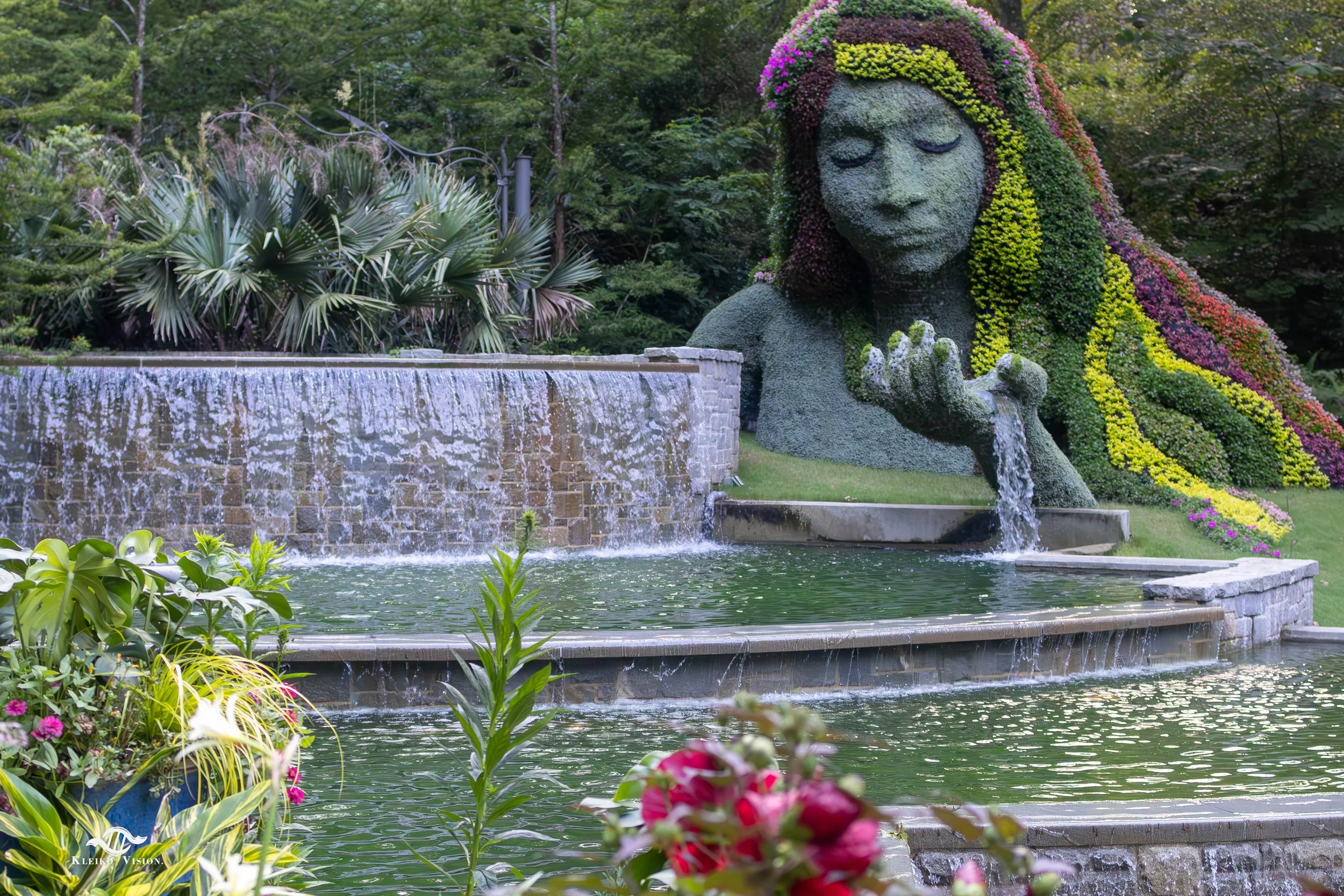 Giant living-plant sculpture of a woman's face pouring water from her hand into a garden pool