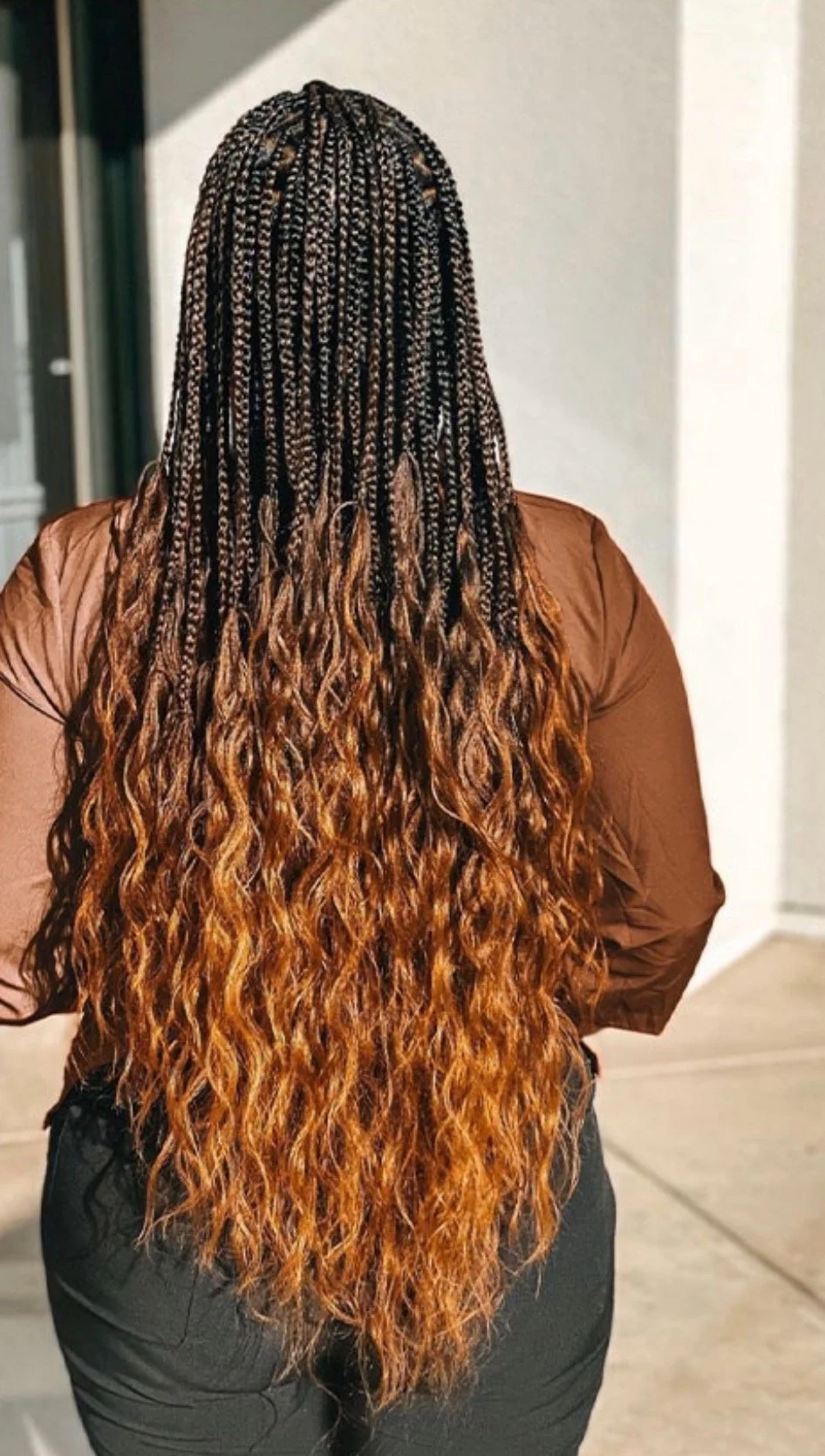 Person with long, curly, brown-to-ginger ombré hair, wearing a brown top and black pants, standing outdoors.