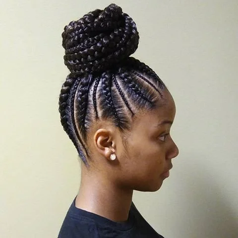 A woman with intricately braided hairstyle styled into a high bun against a neutral wall.