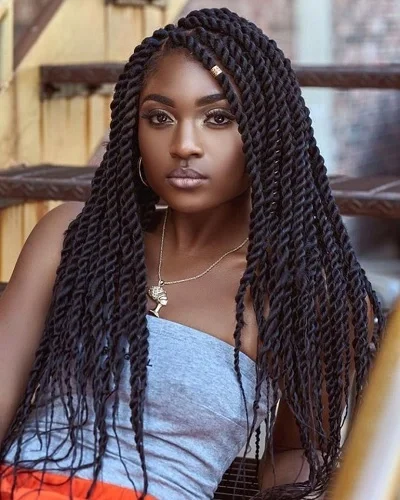 A young woman with long, twisted braids, wearing a gray top and jewelry, sitting outdoors with a serious expression.
