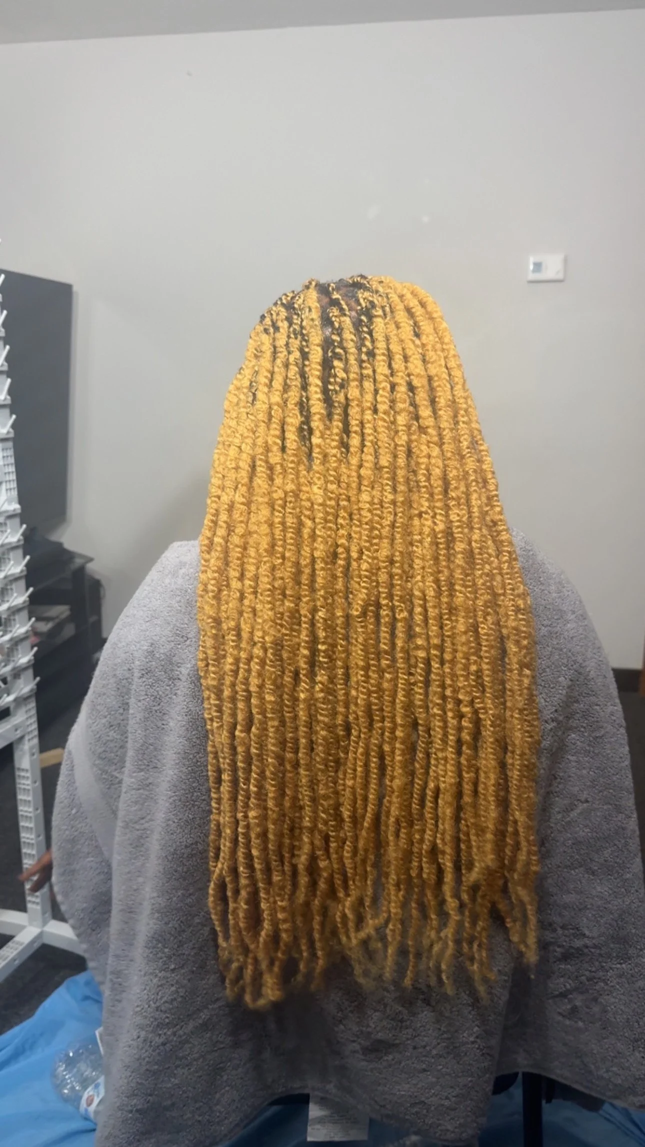 Back view of a person with long blonde locs sitting in a chair, wearing a gray garment in an indoor setting.