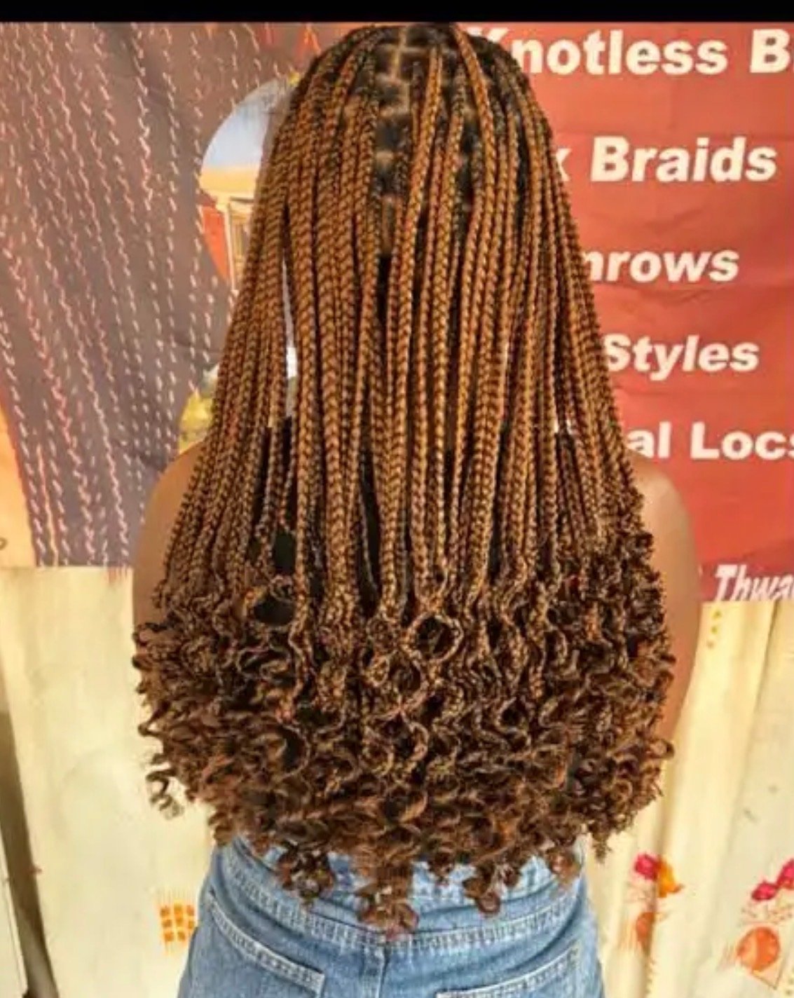 Back view of a woman with long, curly, caramel-colored box braids, standing in front of a red and yellow decor backdrop.