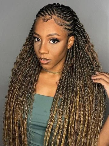 A woman with long, curly, faux locs hairstyle, wearing a green top and a gold necklace, and posing against a plain gray background.