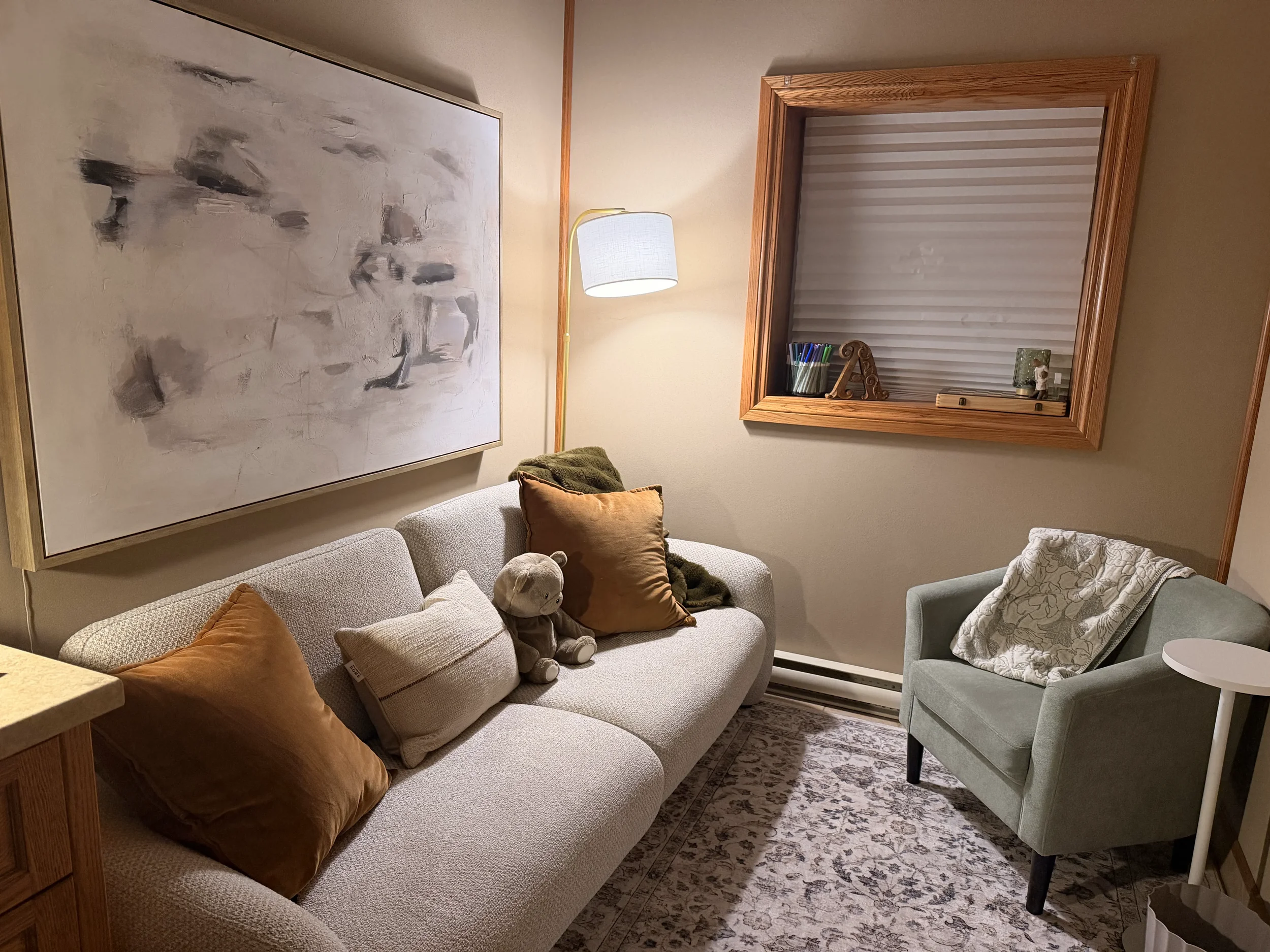 Living room with a beige couch, gray throw blanket, decorative pillow, wooden coffee table with tissue box, white shelving unit with plants and decor, wall clock, framed floral art, and a lamp.
