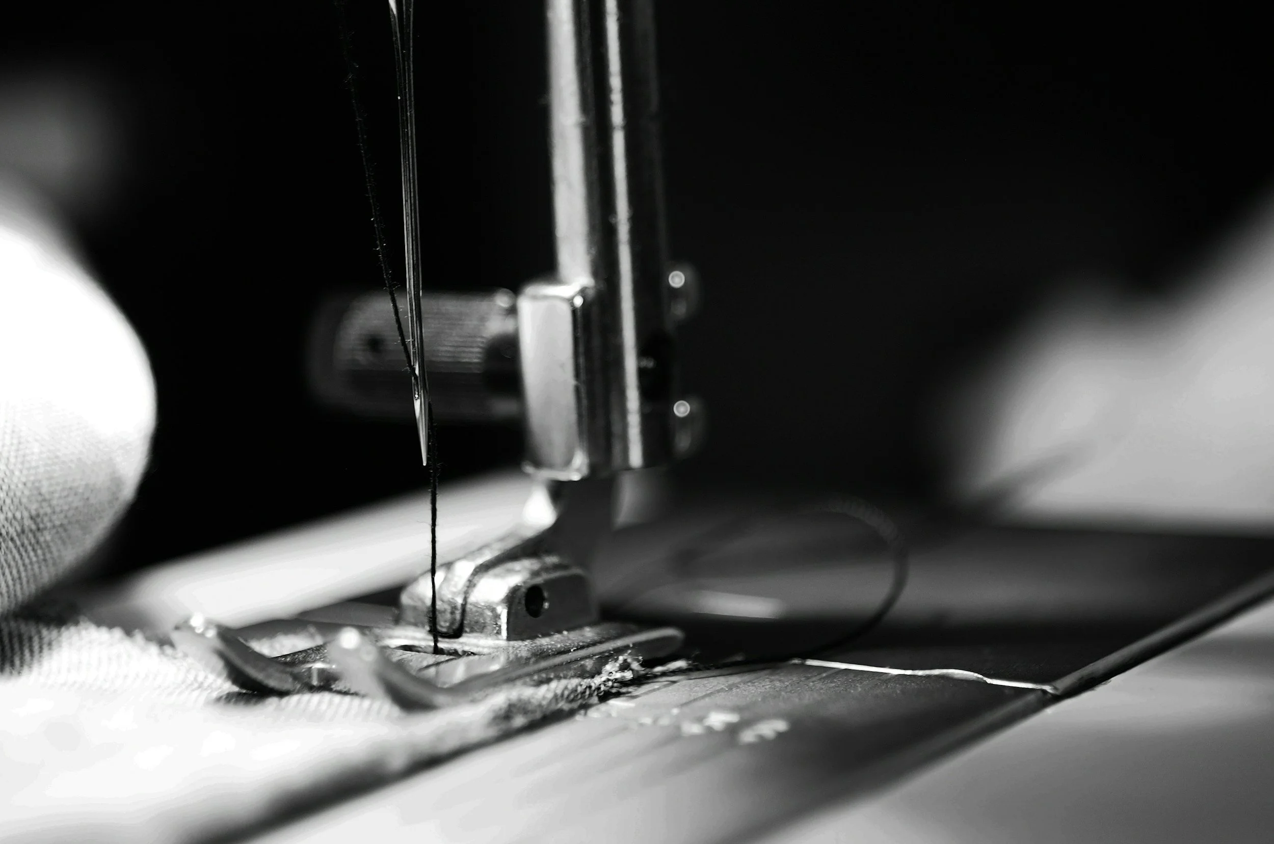 Close-up of a sewing machine needle with black thread moving through metal parts, with fabric underneath.