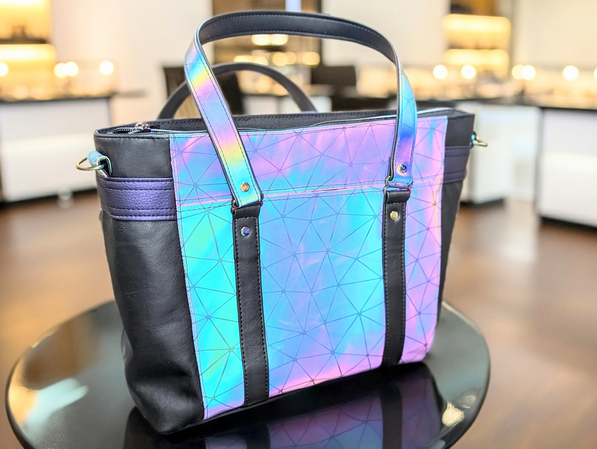 A black tote bag with iridescent panels displaying geometric patterns, placed on a black circular table inside a well-lit store.