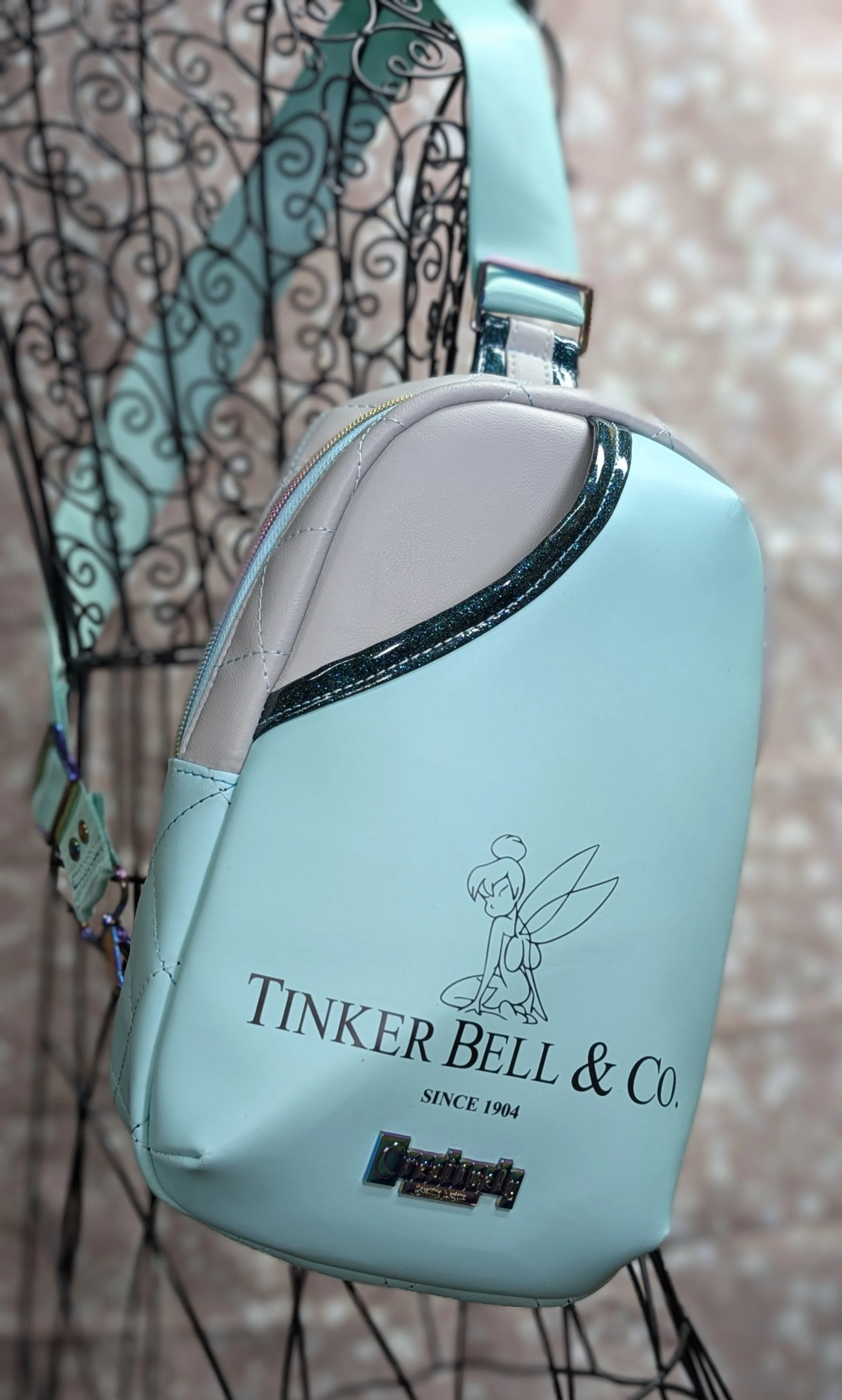Light blue backpack with a fairy illustration and the words "Tinker Bell & Co. Since 1904" on it, hanging from a wire display, with a decorative black iron background.
