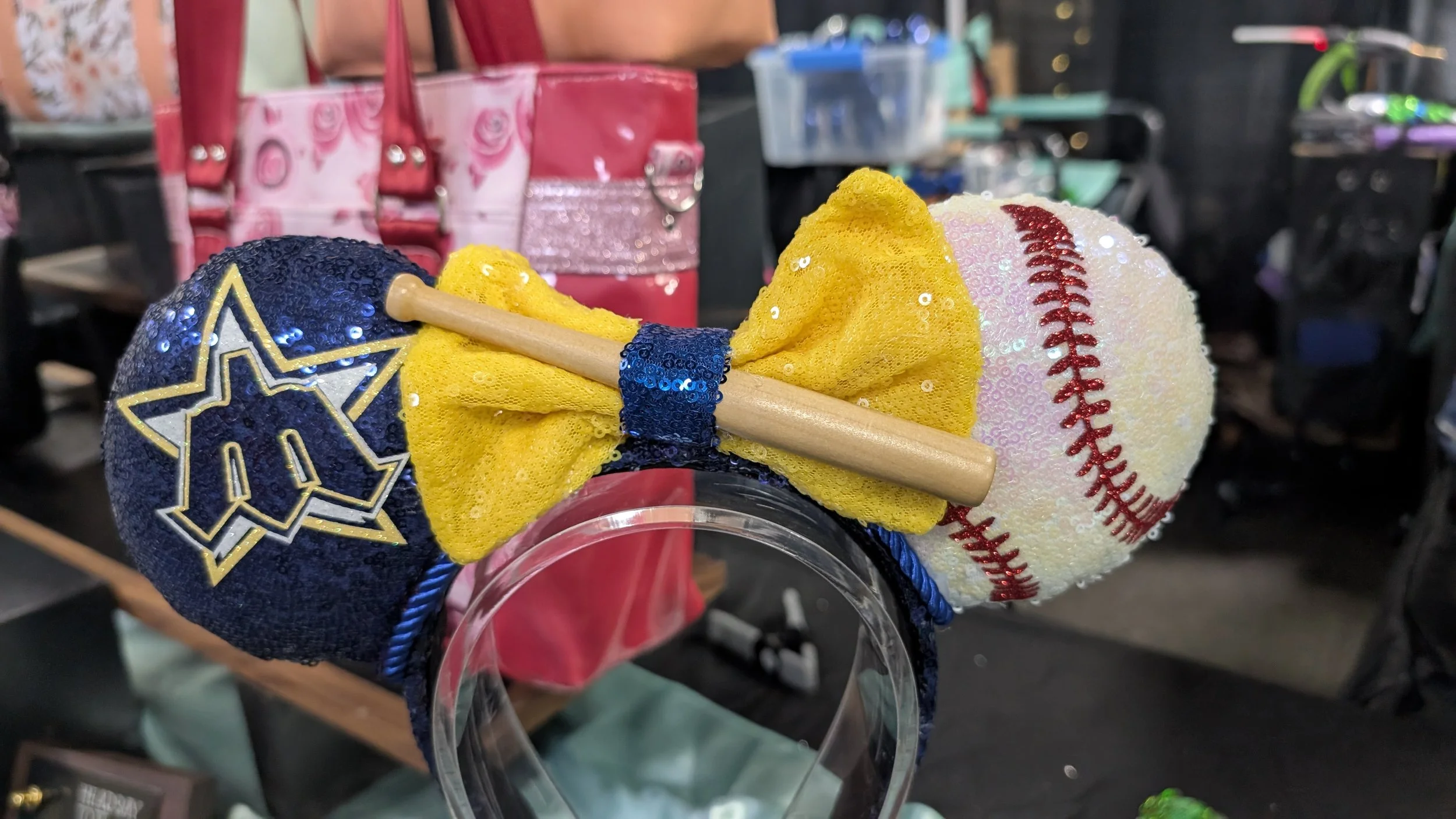 A plush toy designed as a baseball with a baseball hat, bow tie, and a small baseball bat attached. The hat is blue with a star logo, the bow tie is yellow with sequins, and the baseball is white with red stitching. The toy is mounted on a clear plas