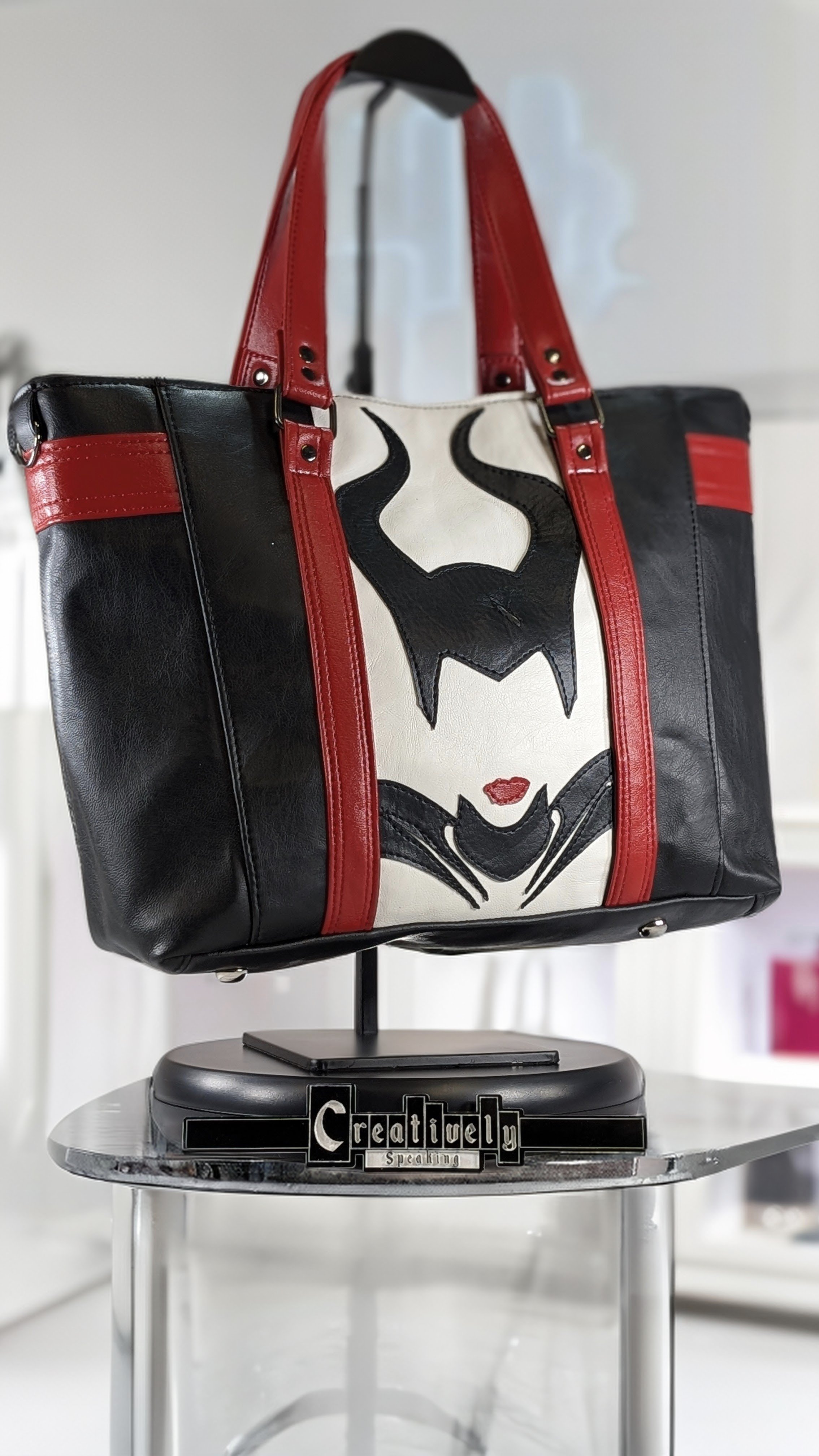 Black tote bag with red straps and a graphic of a woman's face with a bat symbol as hair, red lips, and black cape, displayed on a stand on a table.