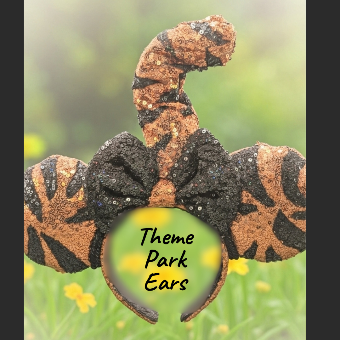 A caterpillar with the appearance of Mickey Mouse ears, covered in black and orange patterns with yellow and shiny spots, on a blurred green and yellow background. The text 'Theme Park Ears' is written on a yellow-green circle at the bottom.
