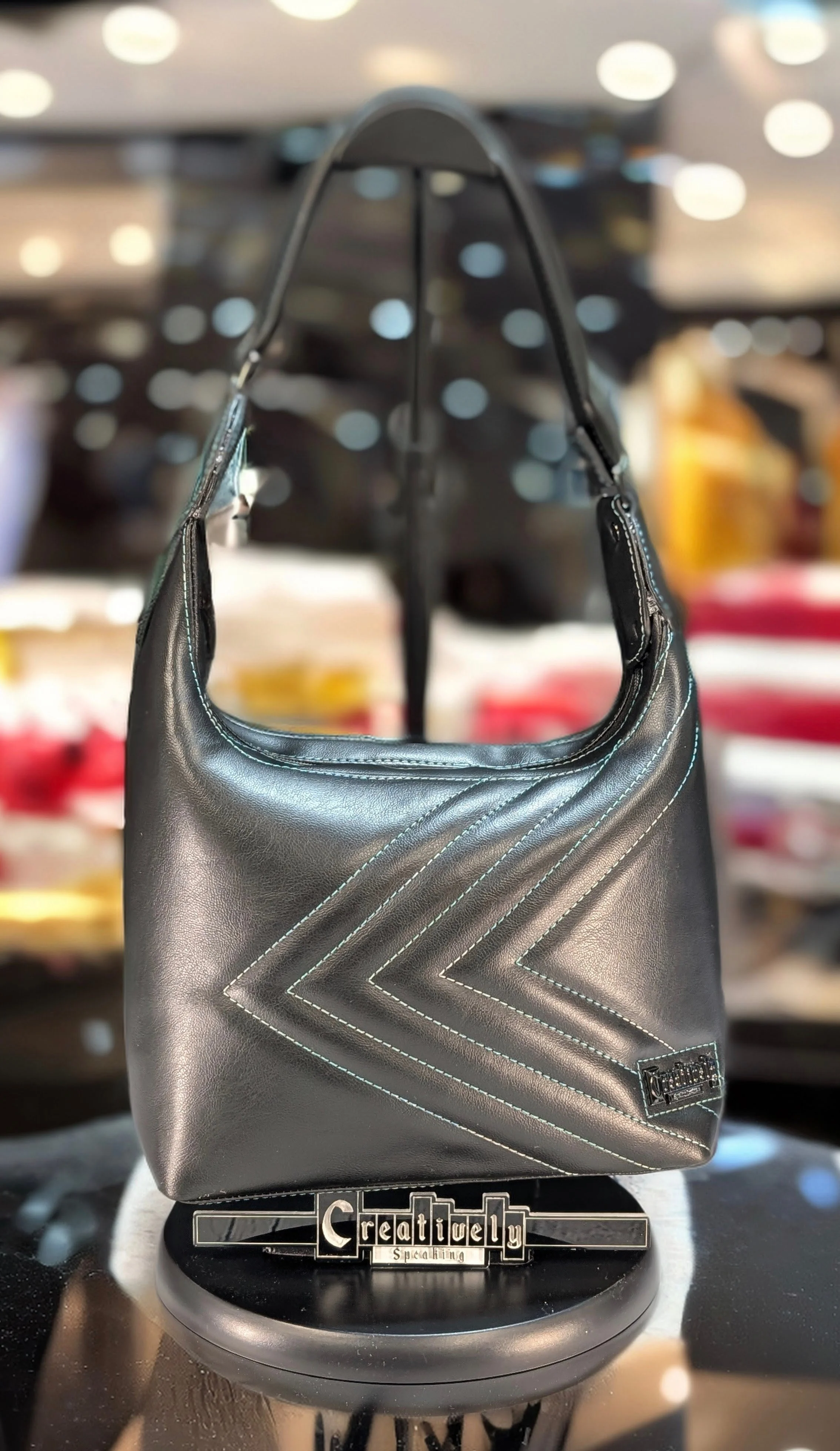 Black leather handbag with white stitched chevron pattern displayed on a table with a black and silver sign reading "Creatively Speaking" in front of it, blurred background of store interior.