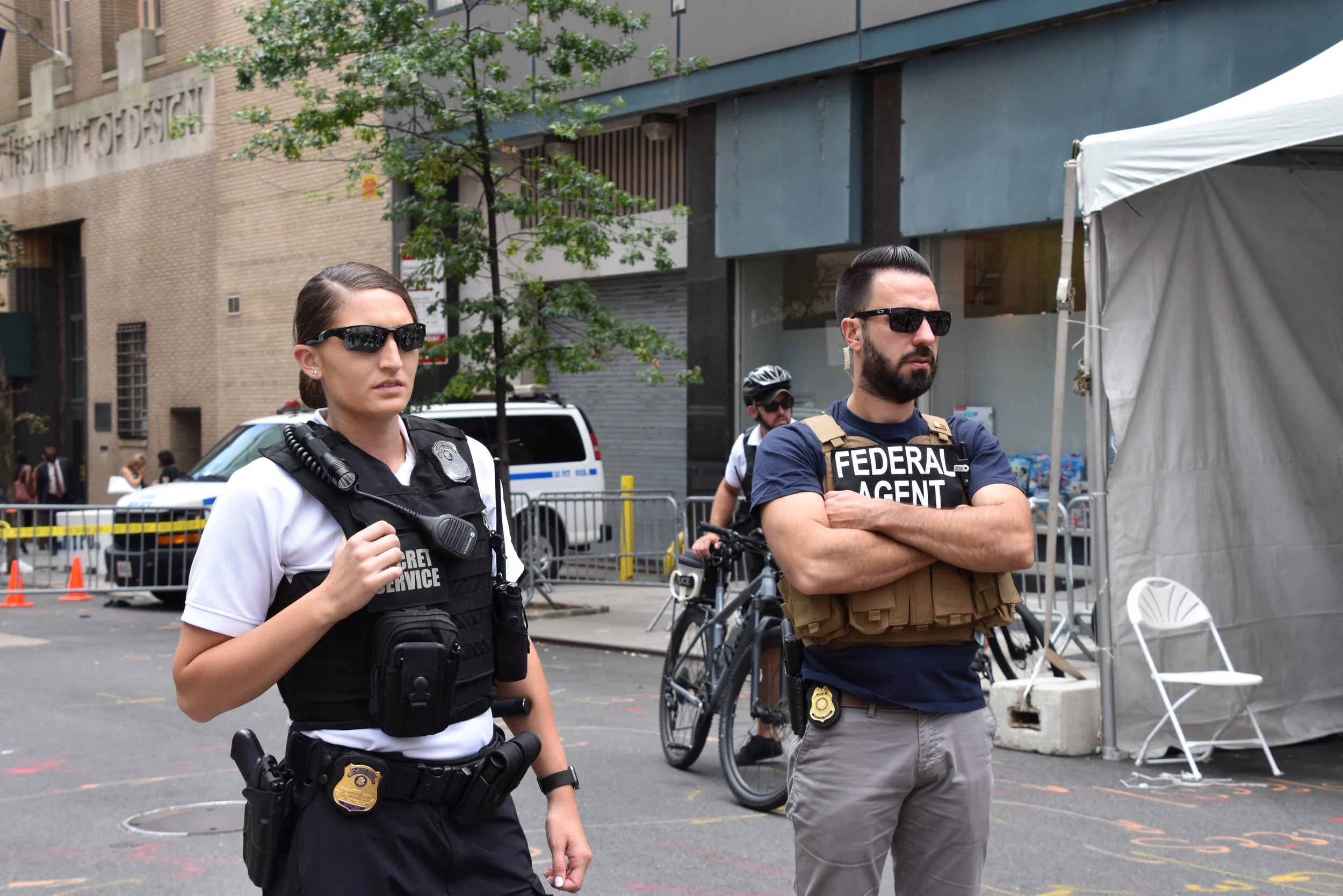 Two law enforcement officers, a female officer and a male officer, are standing outdoors. The female officer is dressed in a police uniform with sunglasses and a radio on her vest. The male officer is wearing a vest labeled 'FEDERAL AGENT' and has his arms crossed. There is a man on a bicycle and a parked police vehicle in the background. A white tent and some orange cones are also visible.
