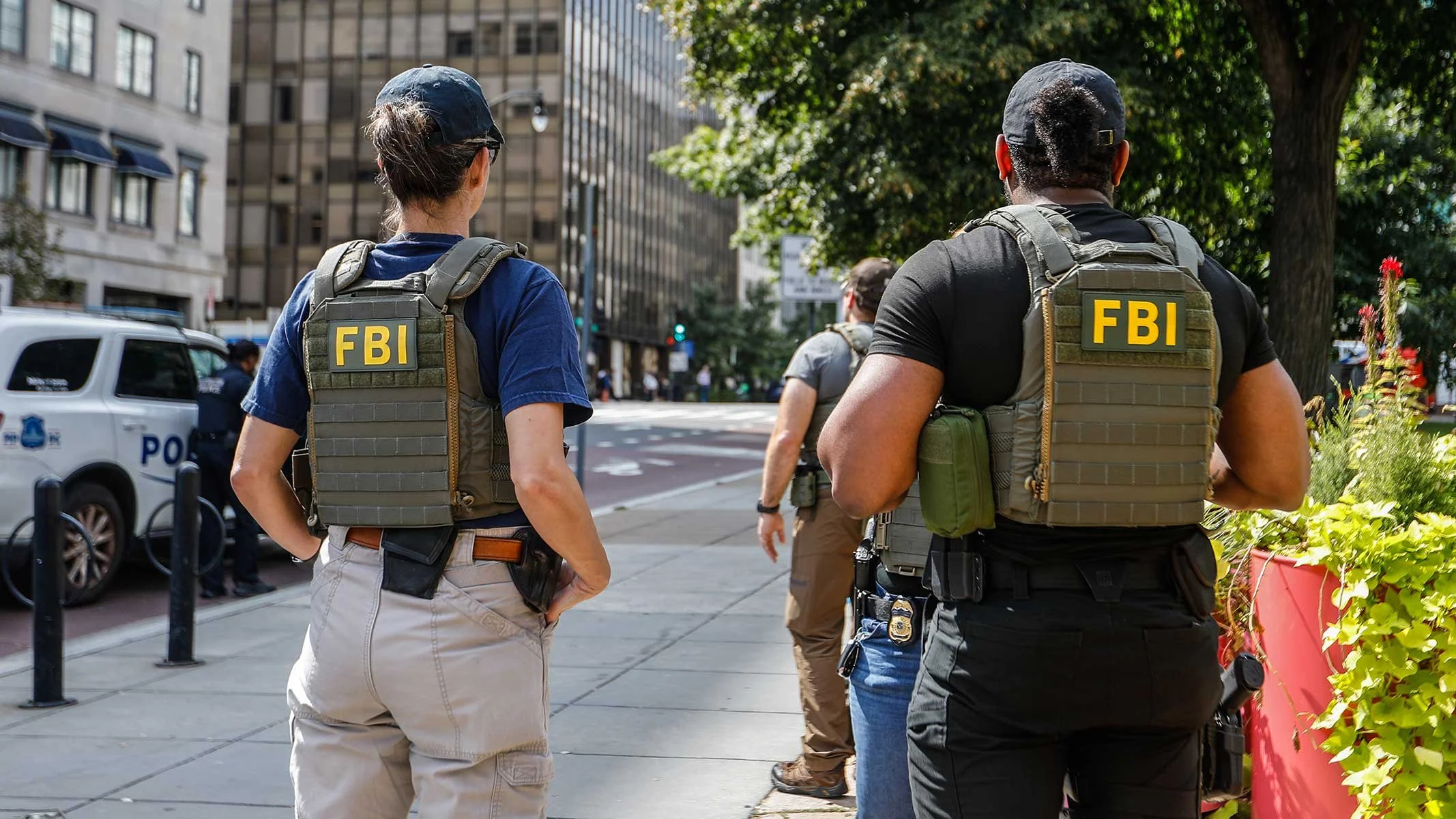 Two FBI agents in tactical gear standing on a city sidewalk, facing away from the camera, with a police car and other people in the background.