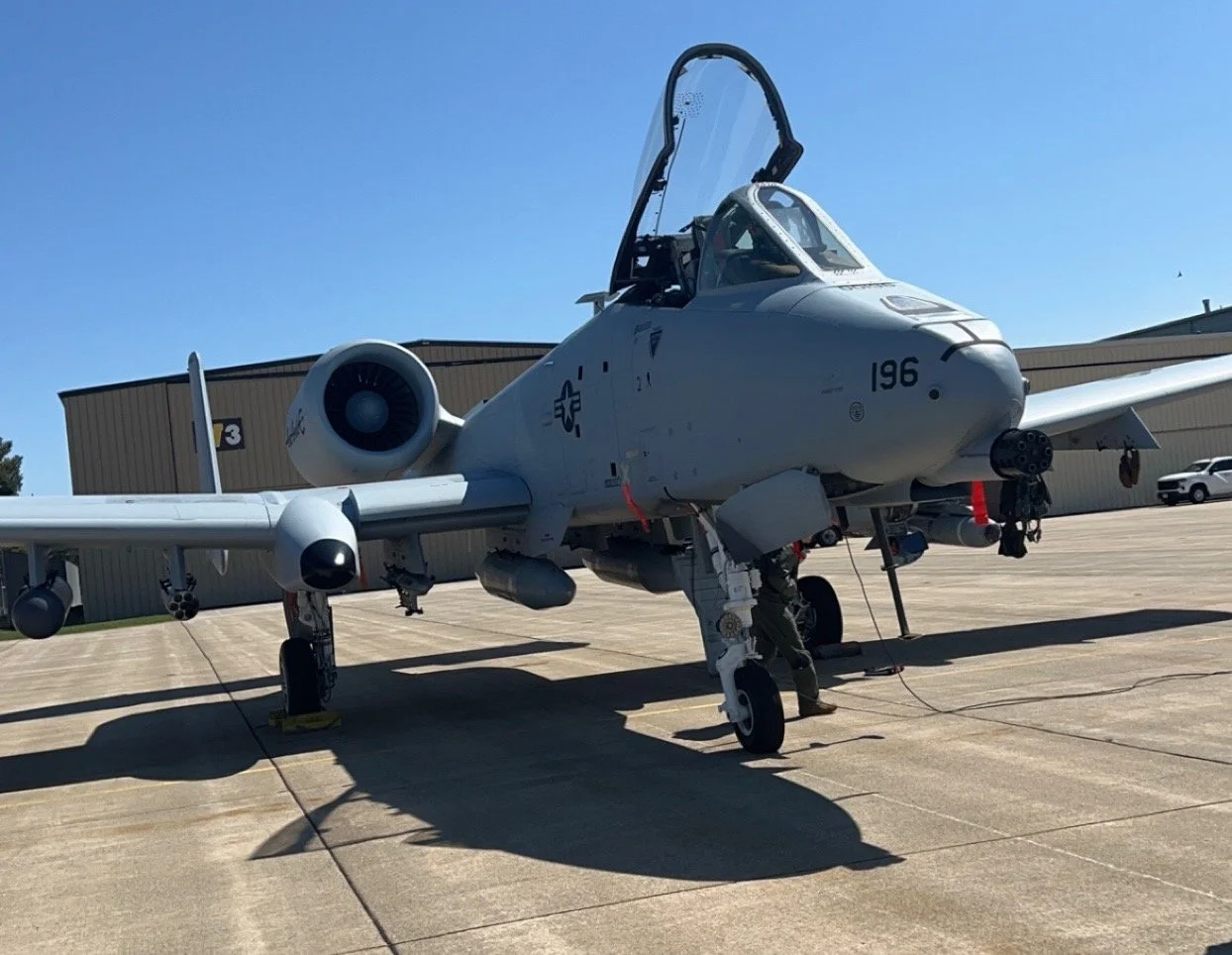A military fighter jet aircraft parked on the tarmac with a person underneath working on it, against a clear blue sky.