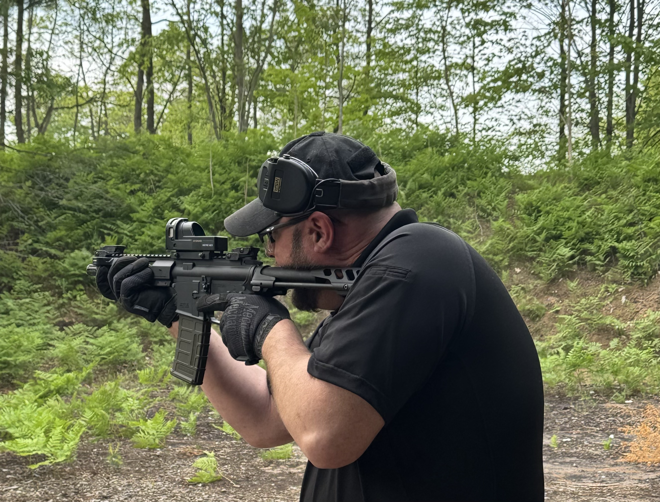 Man wearing black clothing, glasses, and ear protection aims a rifle in a forested area.