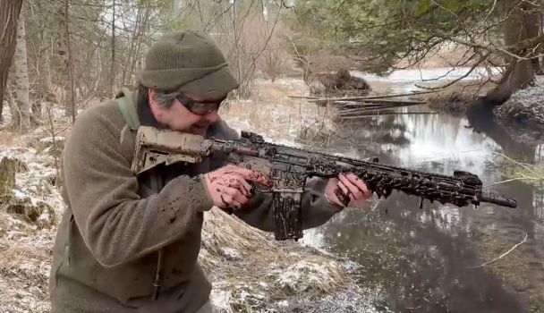 A man outdoors in winter, wearing a brown jacket and a green beanie, aiming a camouflage-patterned sniper rifle with a scope over a small creek.