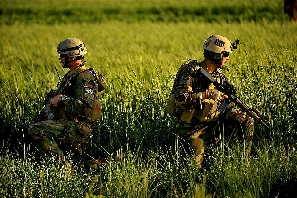 Two soldiers in camouflage uniforms and helmets kneeling in a grassy field, holding rifles and scanning the area.