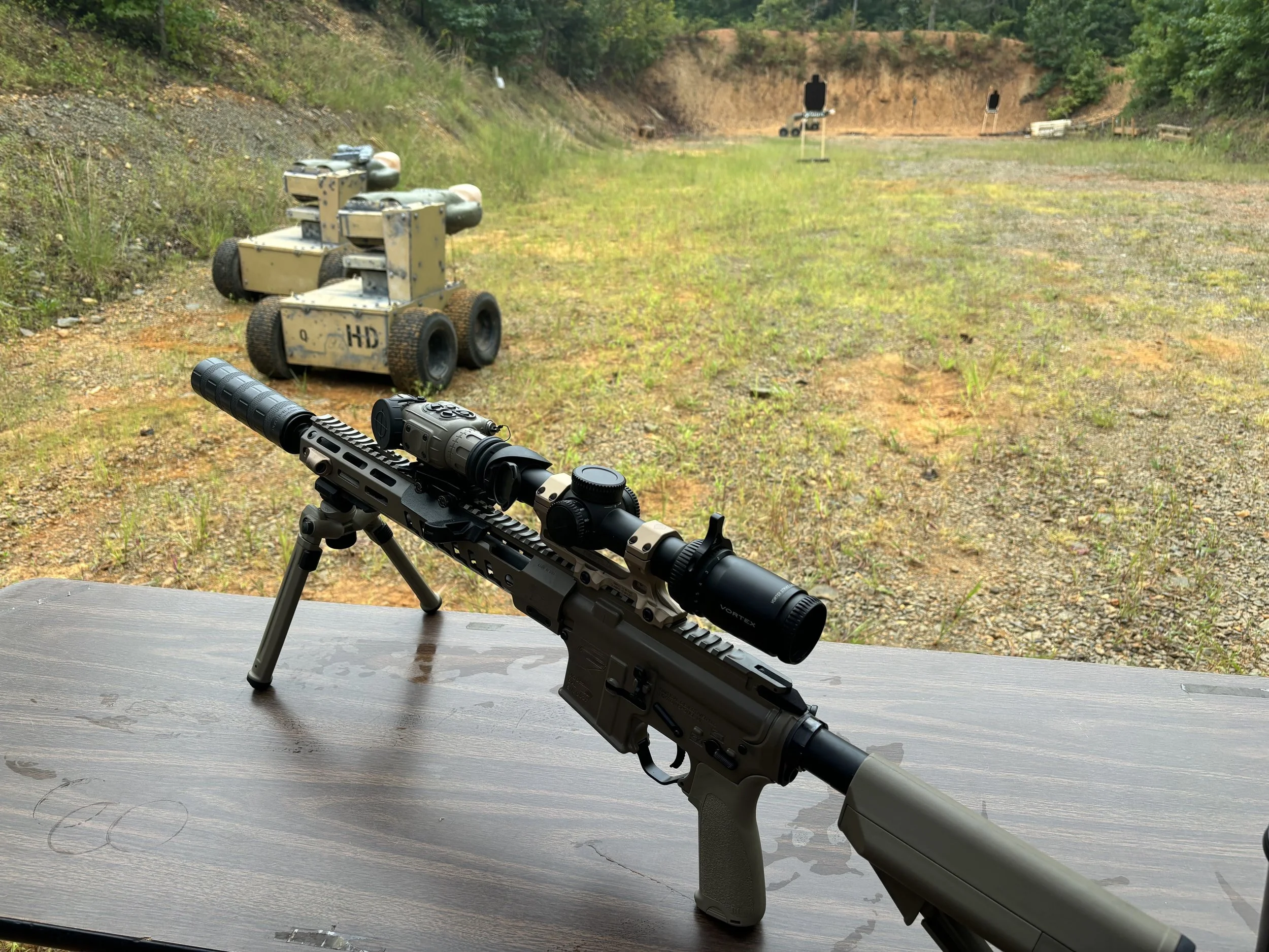A rifle with a scope mounted on a bipod is placed on a wooden table at an outdoor shooting range. In the background, multiple robotic targets and paper targets are set up on a grassy and dirt field with trees behind. The sky is overcast.