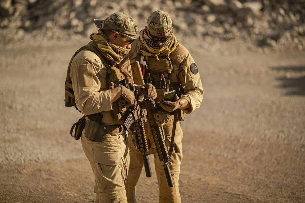 Two soldiers in tan military gear and camouflage hats standing outdoors on a sandy terrain, looking at a handheld device.