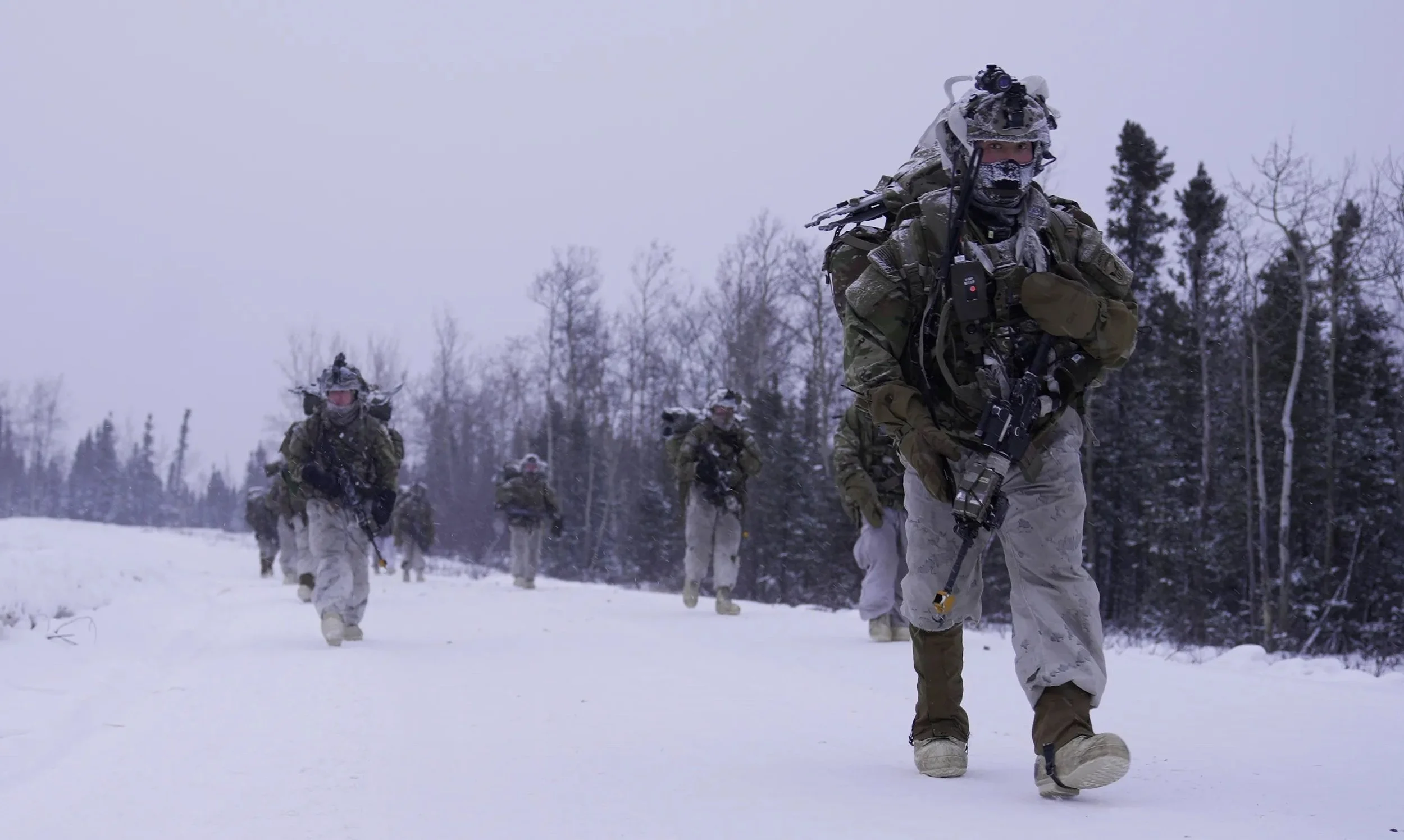 A group of soldiers dressed in winter camouflage, walking through a snowy landscape with trees in the background.