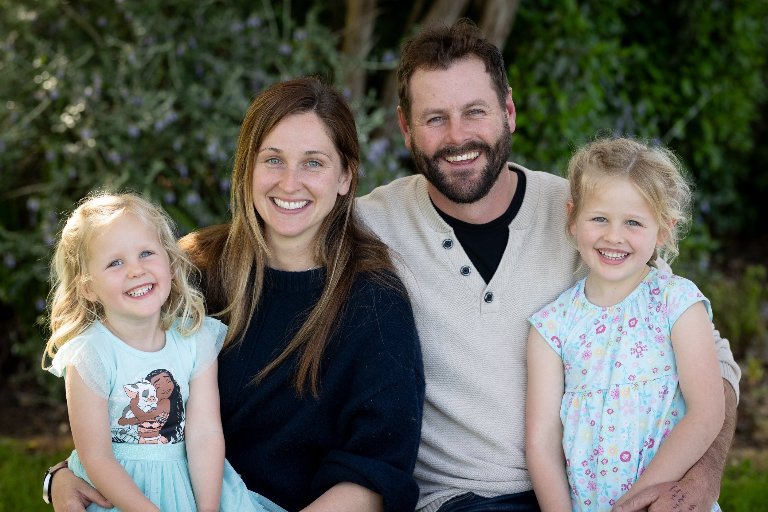 A happy family of four sitting outdoors, with two young girls and two smiling adults, surrounded by greenery.