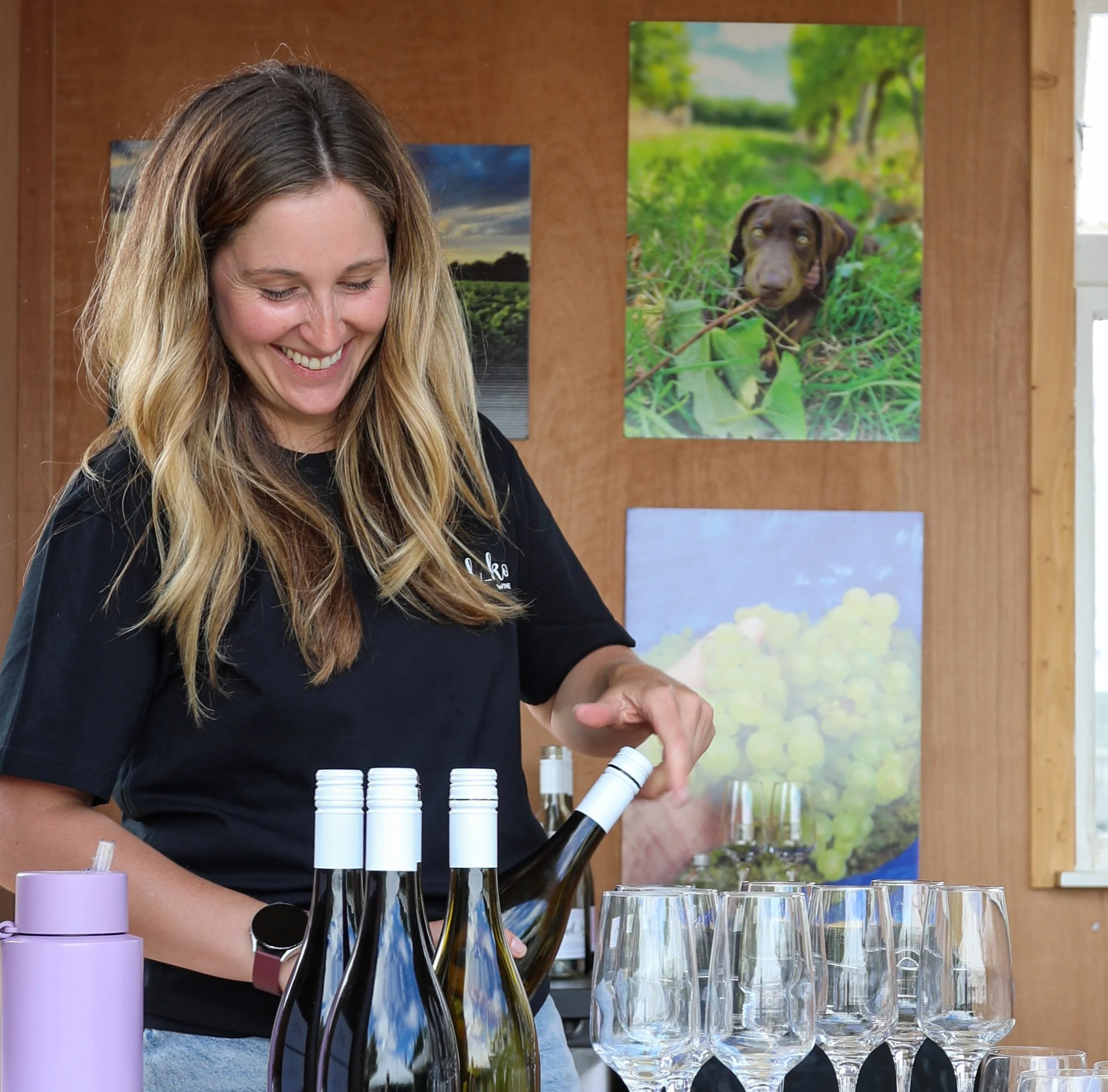 The owner, Hayley is pouring white wine into glasses at their tasting room, with bottles on the table and photos of Arapai vineyard are hanging on the wooden wall behind her.