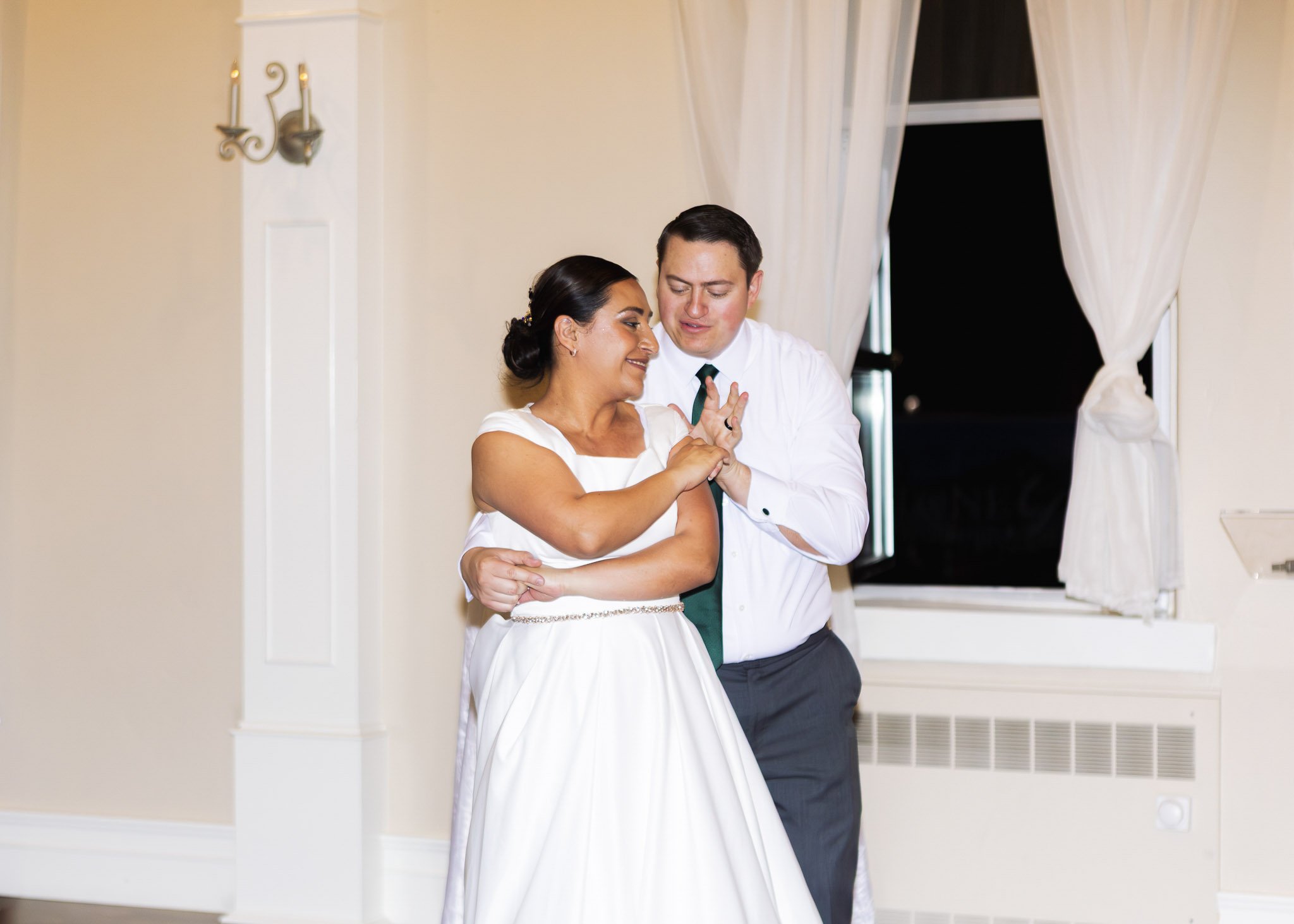A bride and groom sharing a dance indoors at a wedding reception, with white curtains and a window in the background.