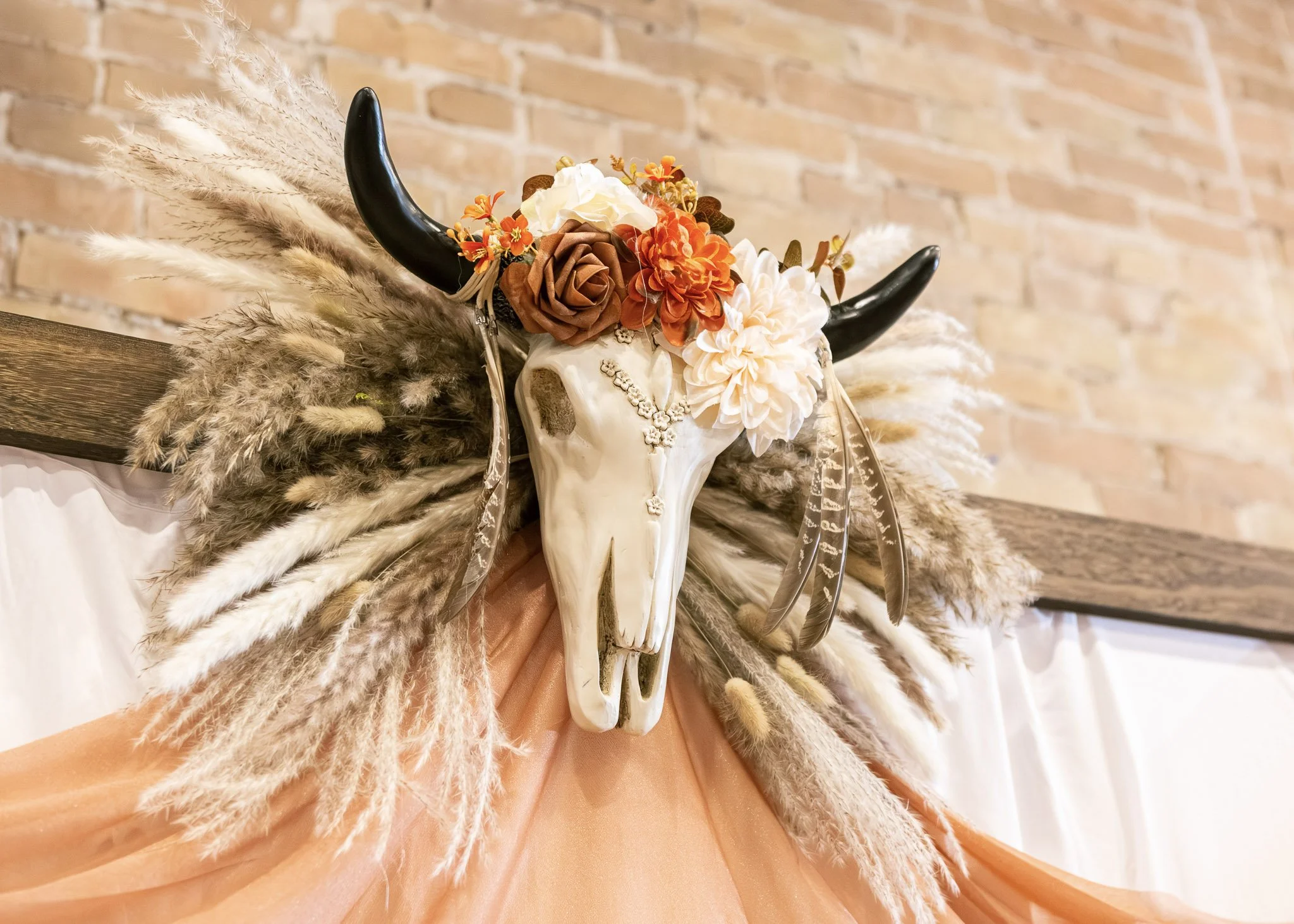 Decorative animal skull with black horns adorned with orange, white, and brown flowers and surrounded by beige pampas grass, set against a brick wall background.
