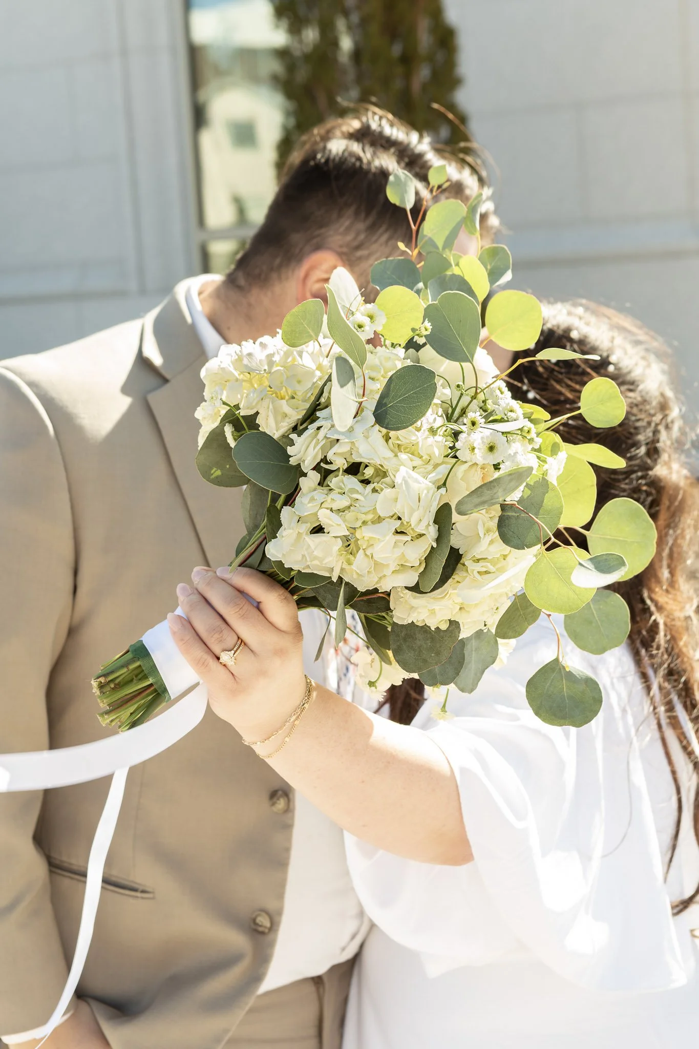 A bride and groom kissing, holding a bouquet of white flowers and green leaves, in front of a window with bright sunlight.