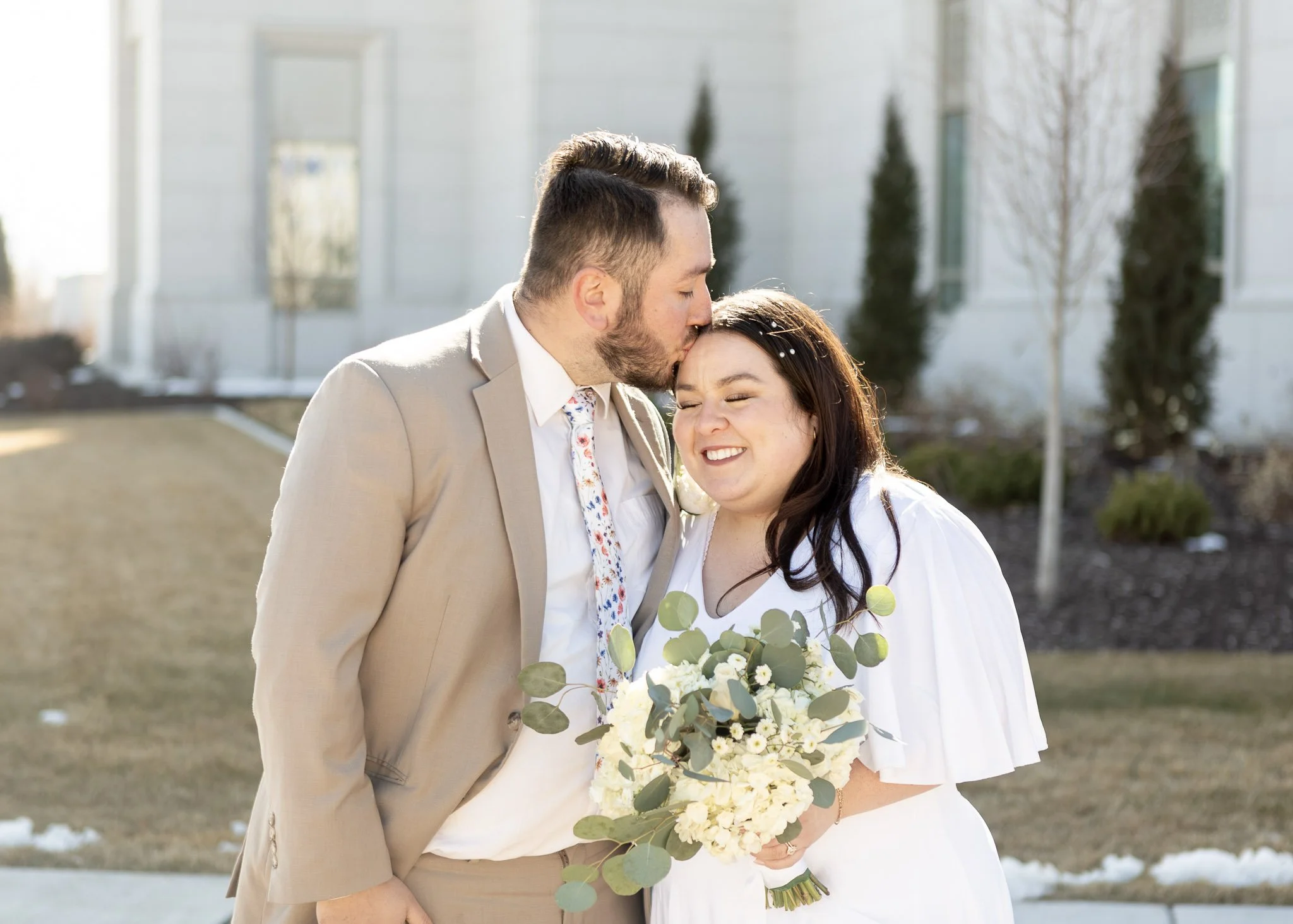 A newly married couple standing outdoors, the man is kissing the woman's forehead, and she is holding a bouquet of white flowers with green leaves.