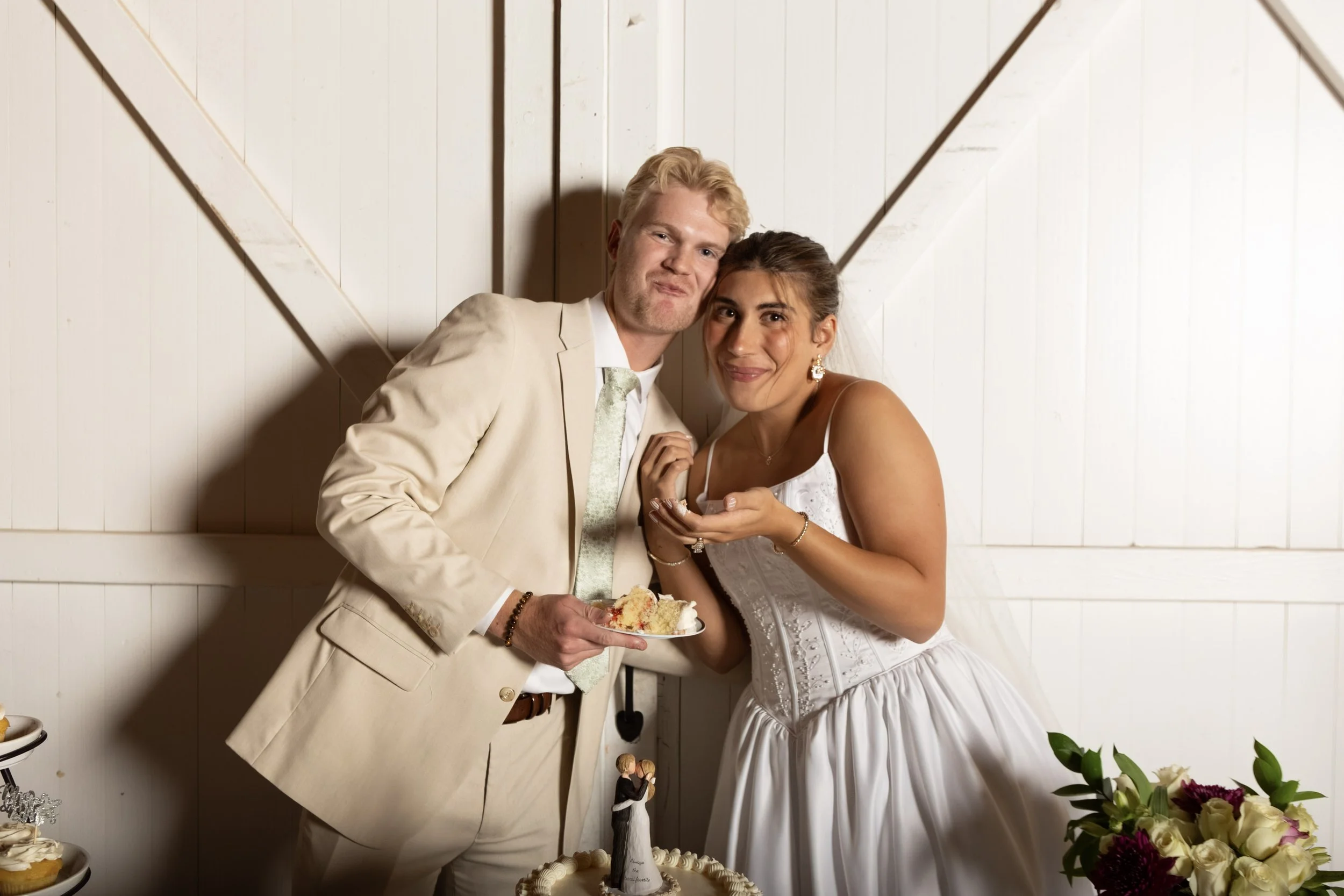 A man and woman in wedding attire stand close together, smiling at the camera. The man holds a piece of cake on a plate, and the woman holds a fork and a piece of cake. There are flowers and a wedding cake with a topper in front of them.