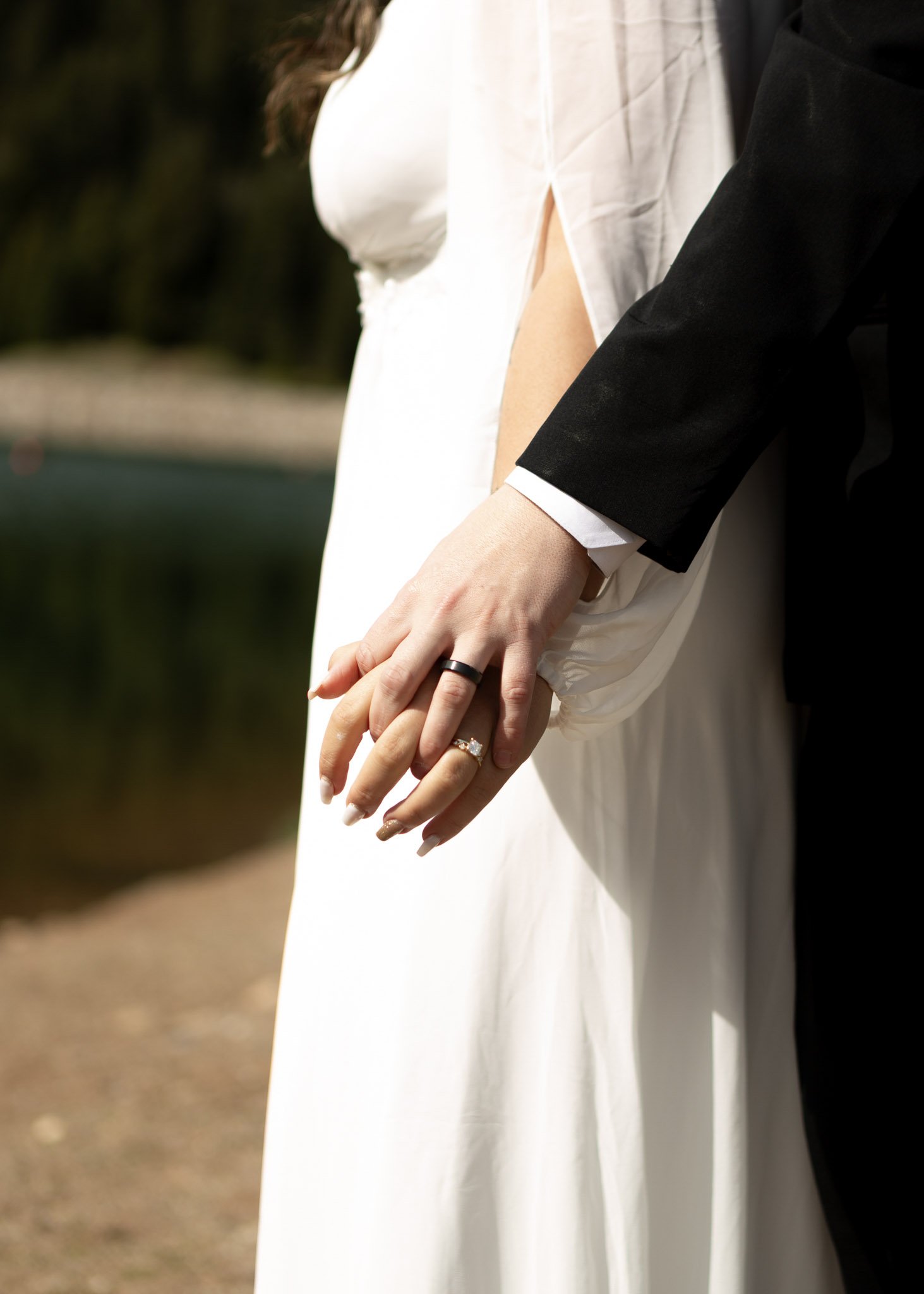 Close-up of a bride and groom holding hands, focusing on their intertwined fingers and wedding rings, with a blurred outdoor background.