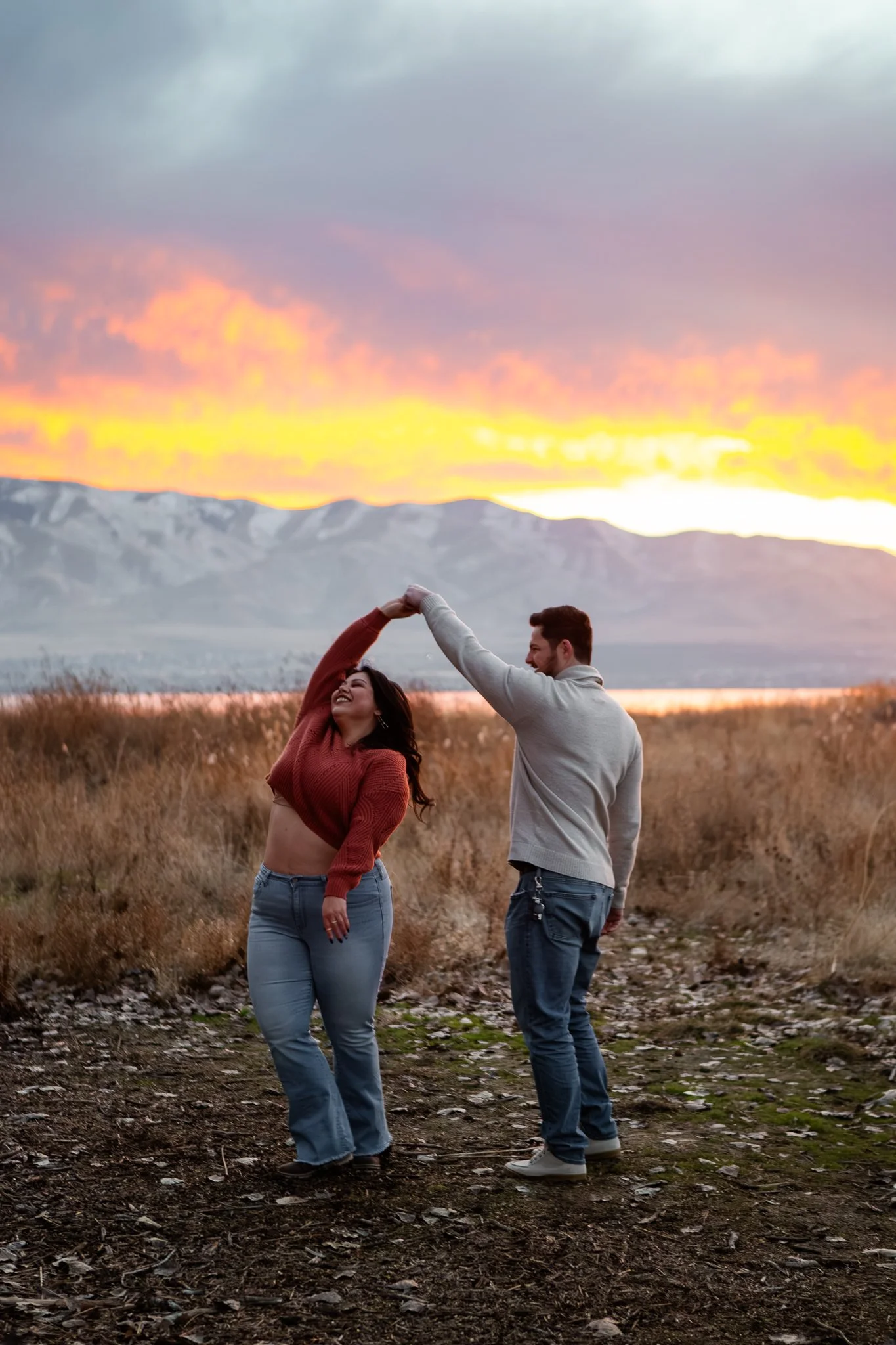 A couple dancing outdoors during a vibrant sunset with mountains in the background, woman in a red sweater and jeans, man in a light hoodie and jeans.