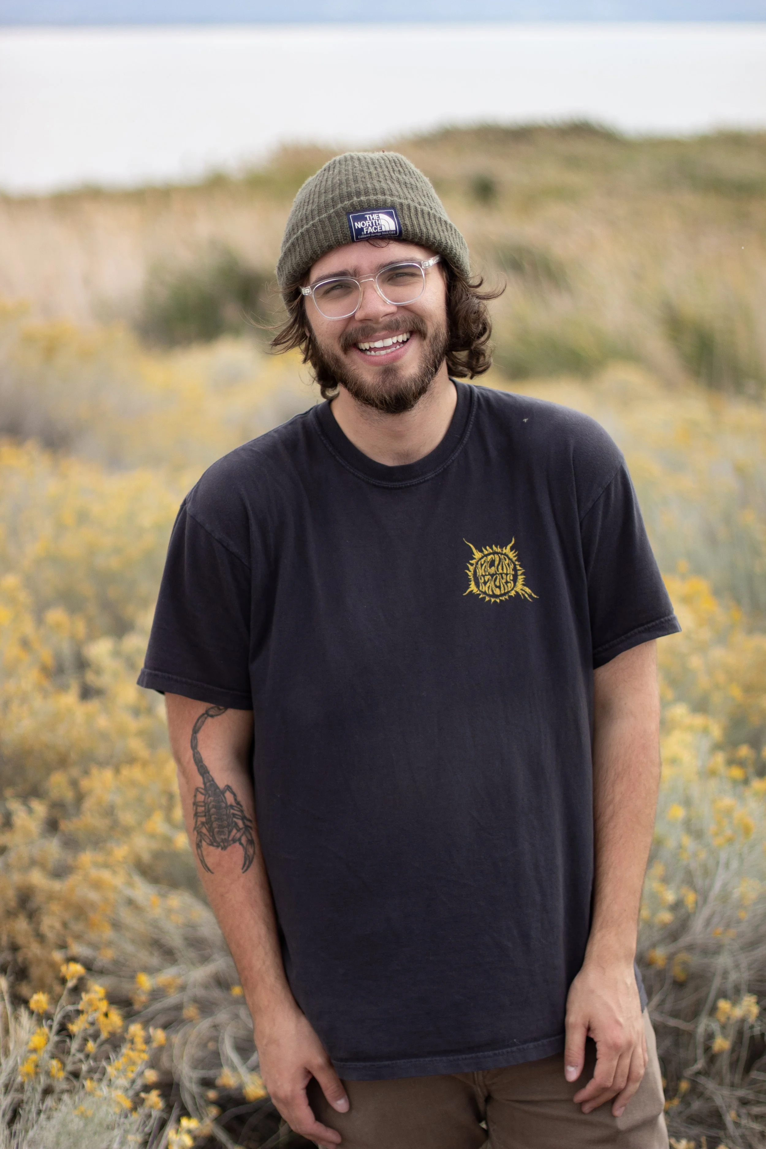 A smiling man with glasses, a camera shy beard, and long hair, wearing a green beanie and a black T-shirt, standing outdoors in a field of yellow wildflowers with a body of water and a hill in the background.