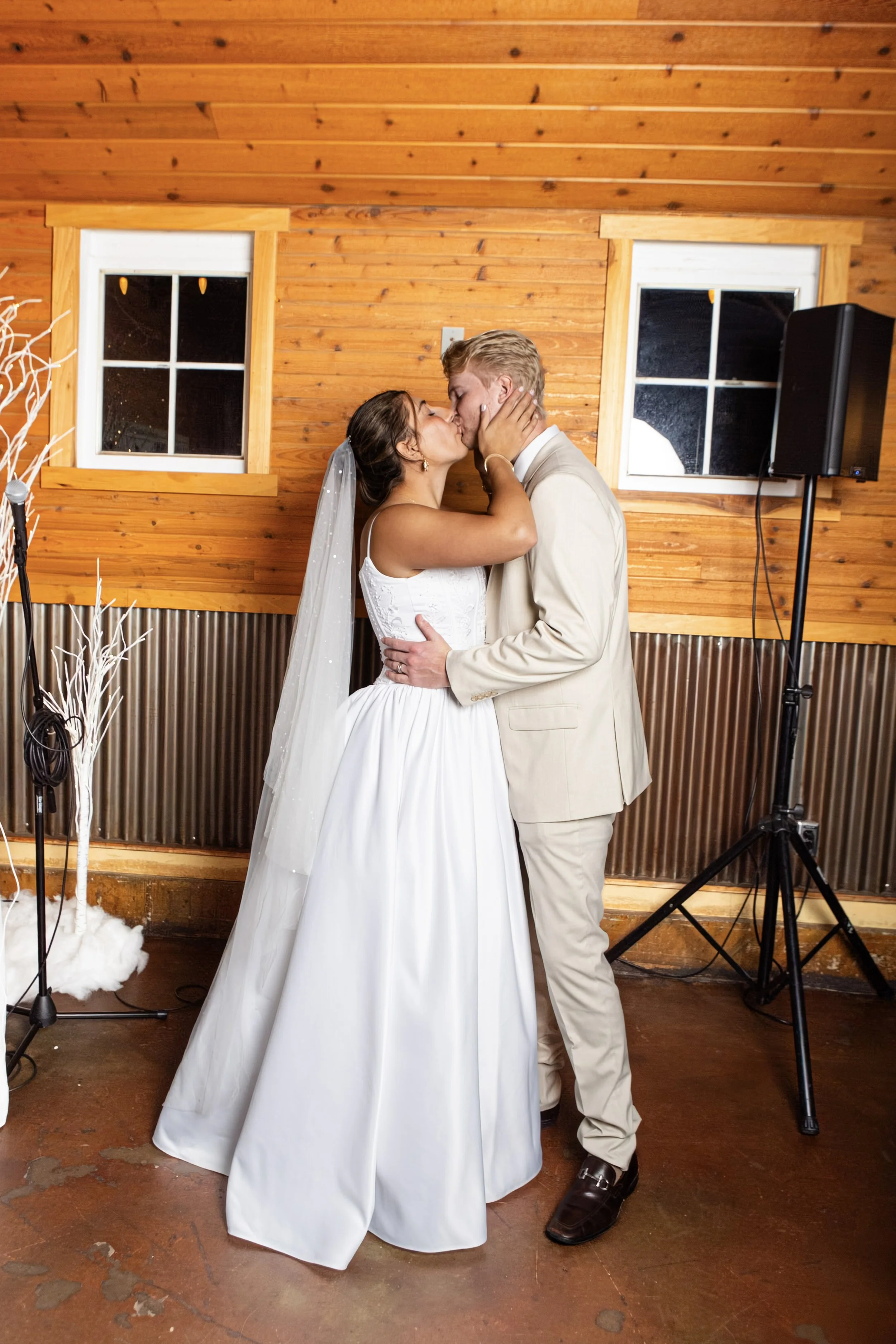 A bride and groom kissing during a wedding reception in a wooden room with two windows and a speaker on a stand.