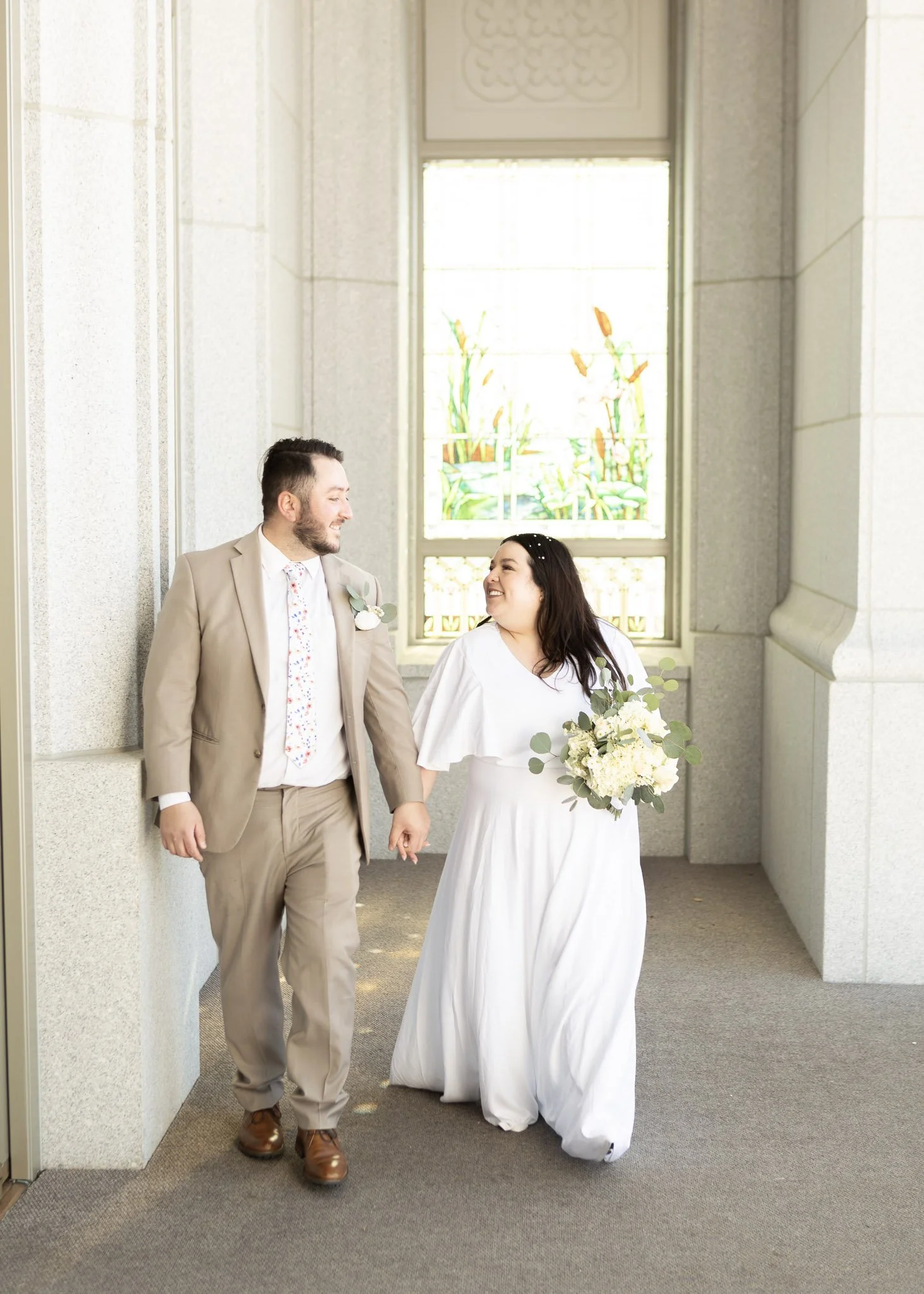 A newly married couple holding hands inside a building with a large window in the background. The man is wearing a beige suit and the woman is wearing a white dress and holding a bouquet of white flowers and greenery. They are smiling and looking at 