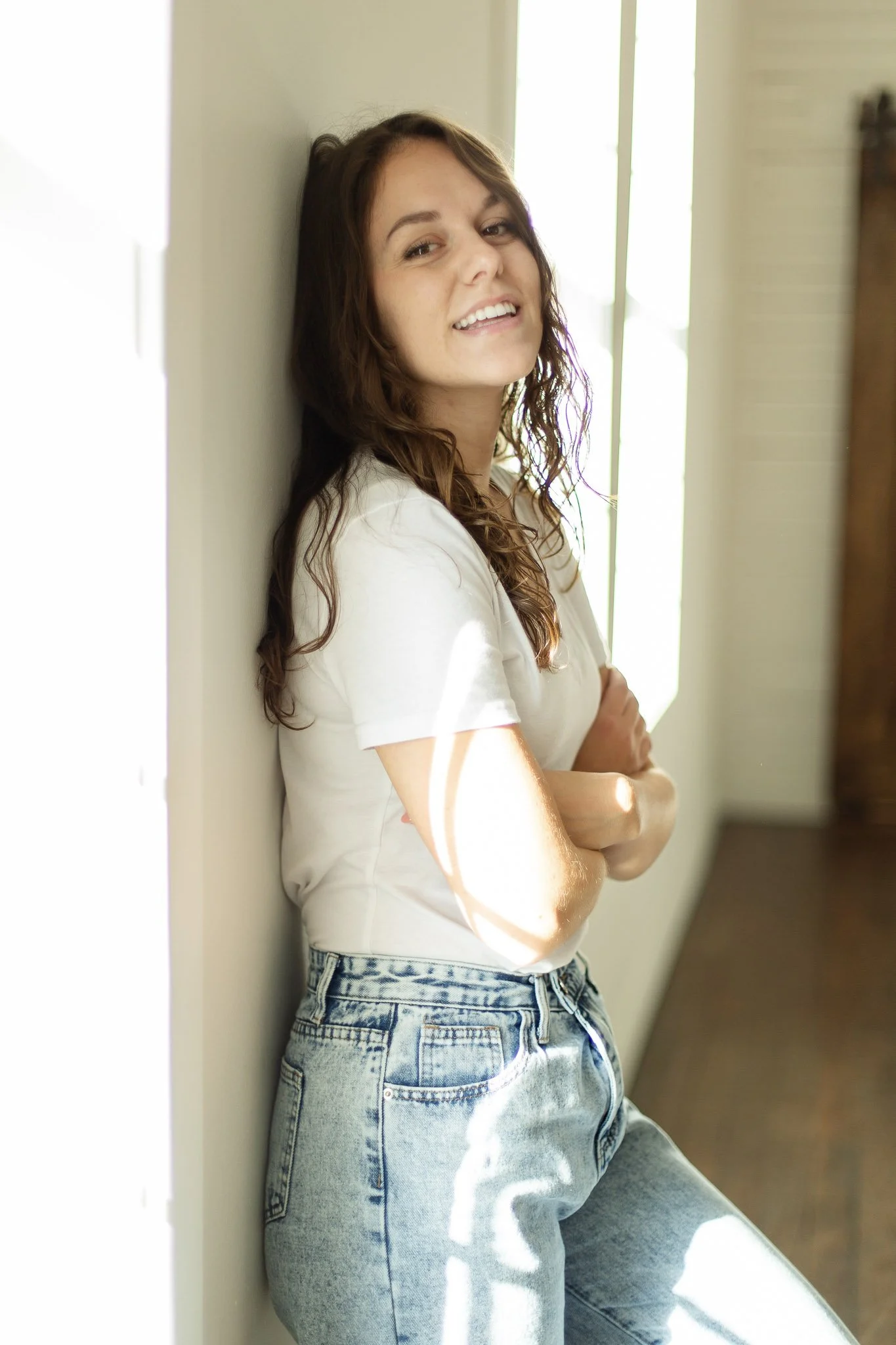 A young woman with wavy brown hair leaning against a wall near a bright window, smiling at the camera, wearing a white t-shirt and high-waisted jeans. Located indoors at a photography studio.